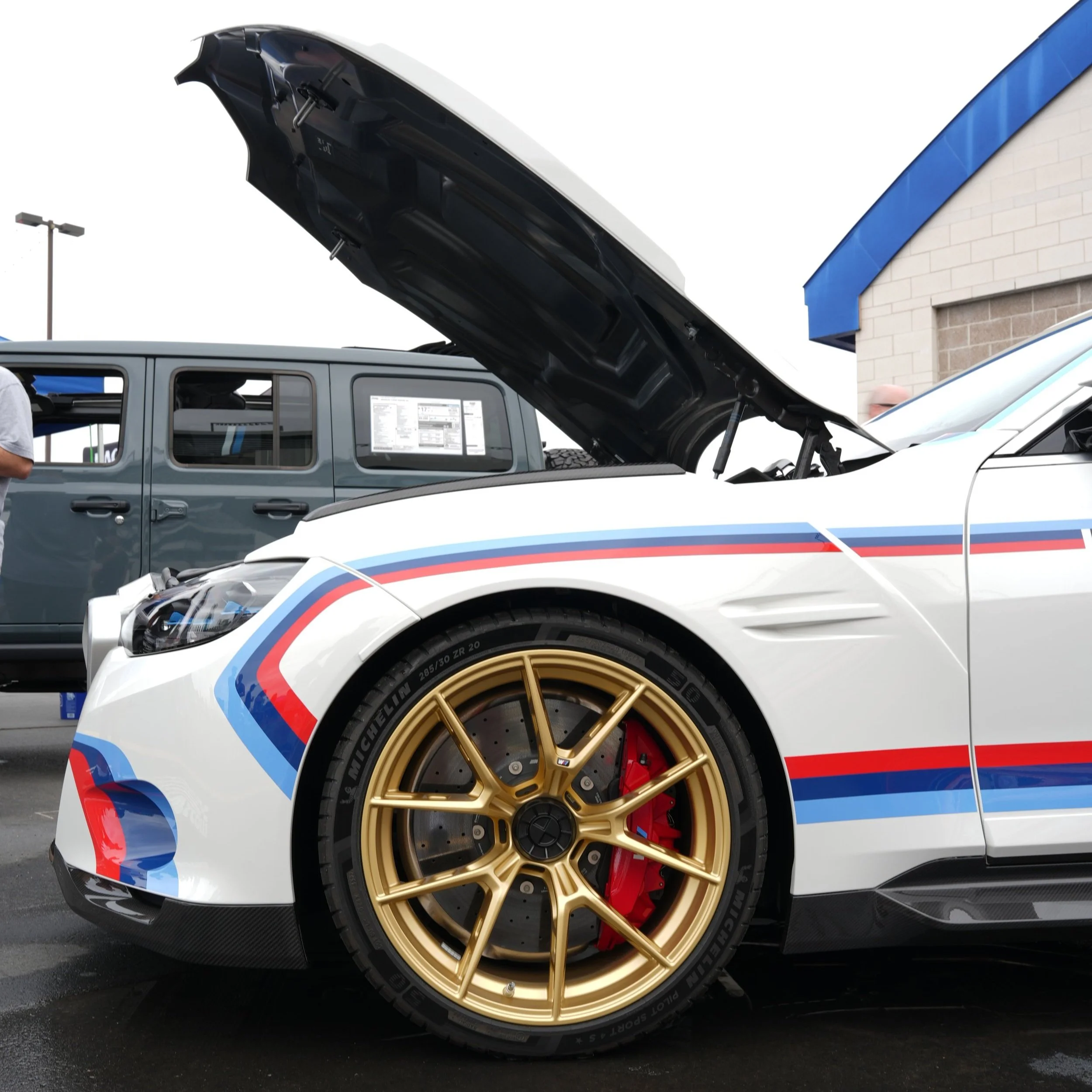 A white sports car with red and blue racing stripes and gold wheels with red brake calipers, parked at a dealership lot with its hood open.