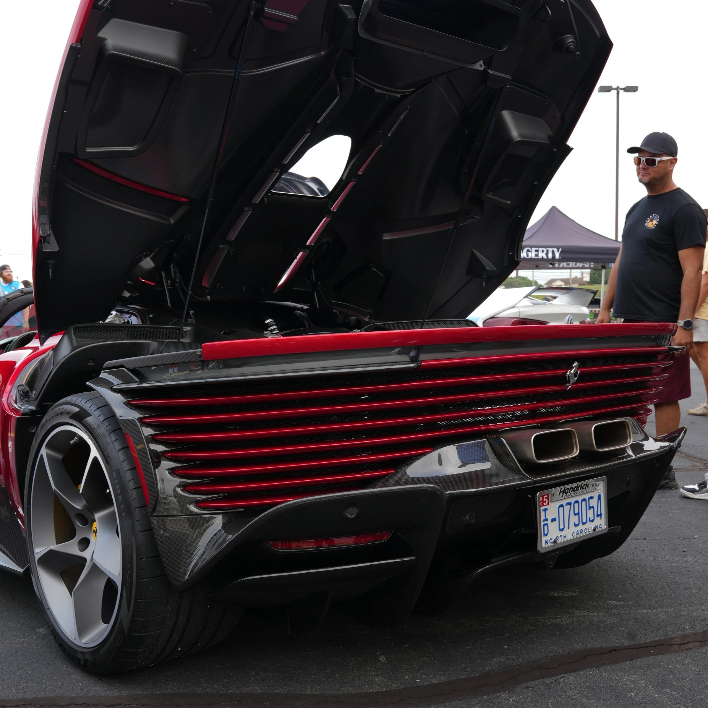 A red and black sports car with its rear engine cover open, showcasing the engine bay. The car has a license plate from North Carolina and is displayed at an automotive event, with a man in a black t-shirt, shorts, sunglasses, and a cap standing near