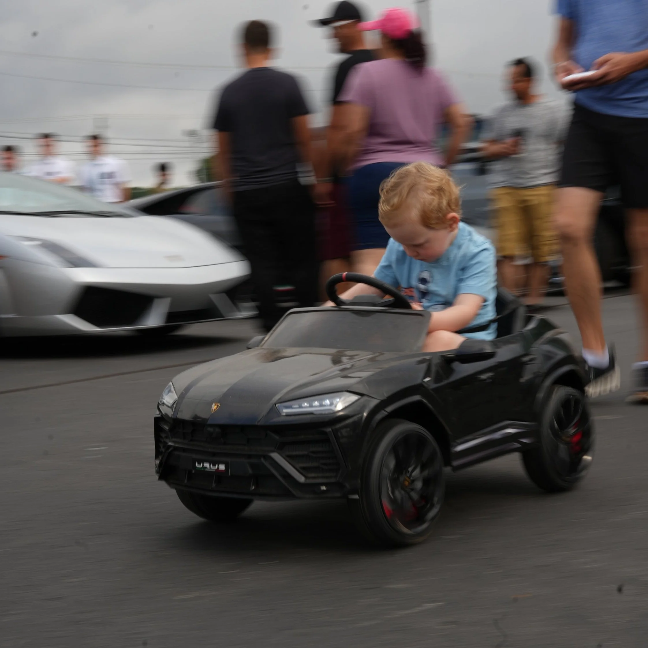 A young boy with red hair is riding a small black toy Lamborghini in a parking lot, with people and luxury cars in the background.
