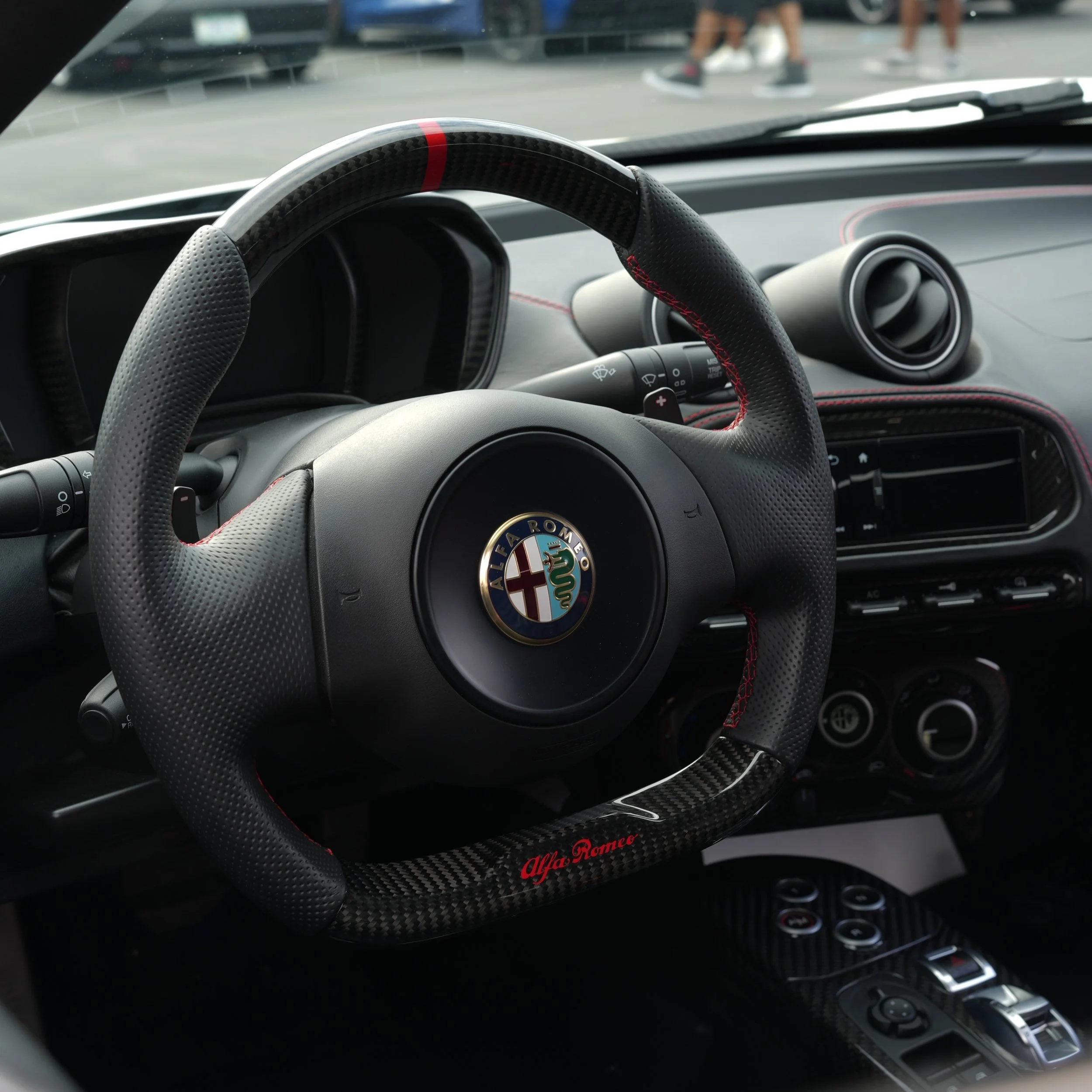 Interior of an Alfa Romeo sports car, showing a carbon fiber steering wheel with the Alfa Romeo logo in the center, and a dashboard with air vents and controls.