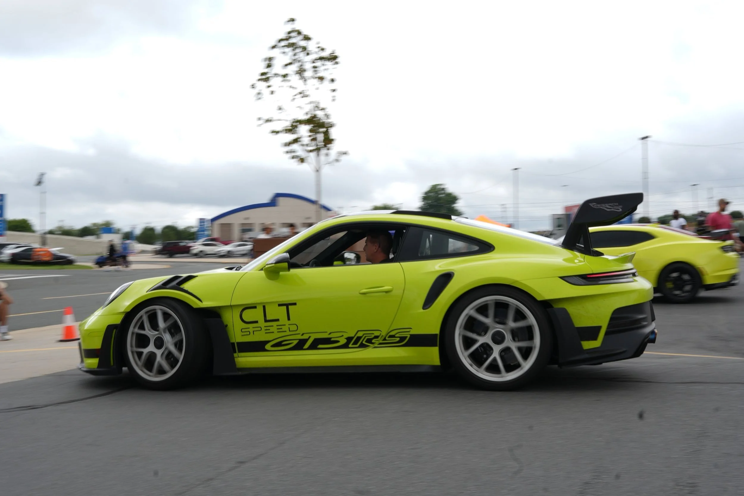A bright yellow Porsche 911 GT3 RS sports car with black decals and a large rear wing, driven on a paved surface during a car event, with other cars and people in the background.