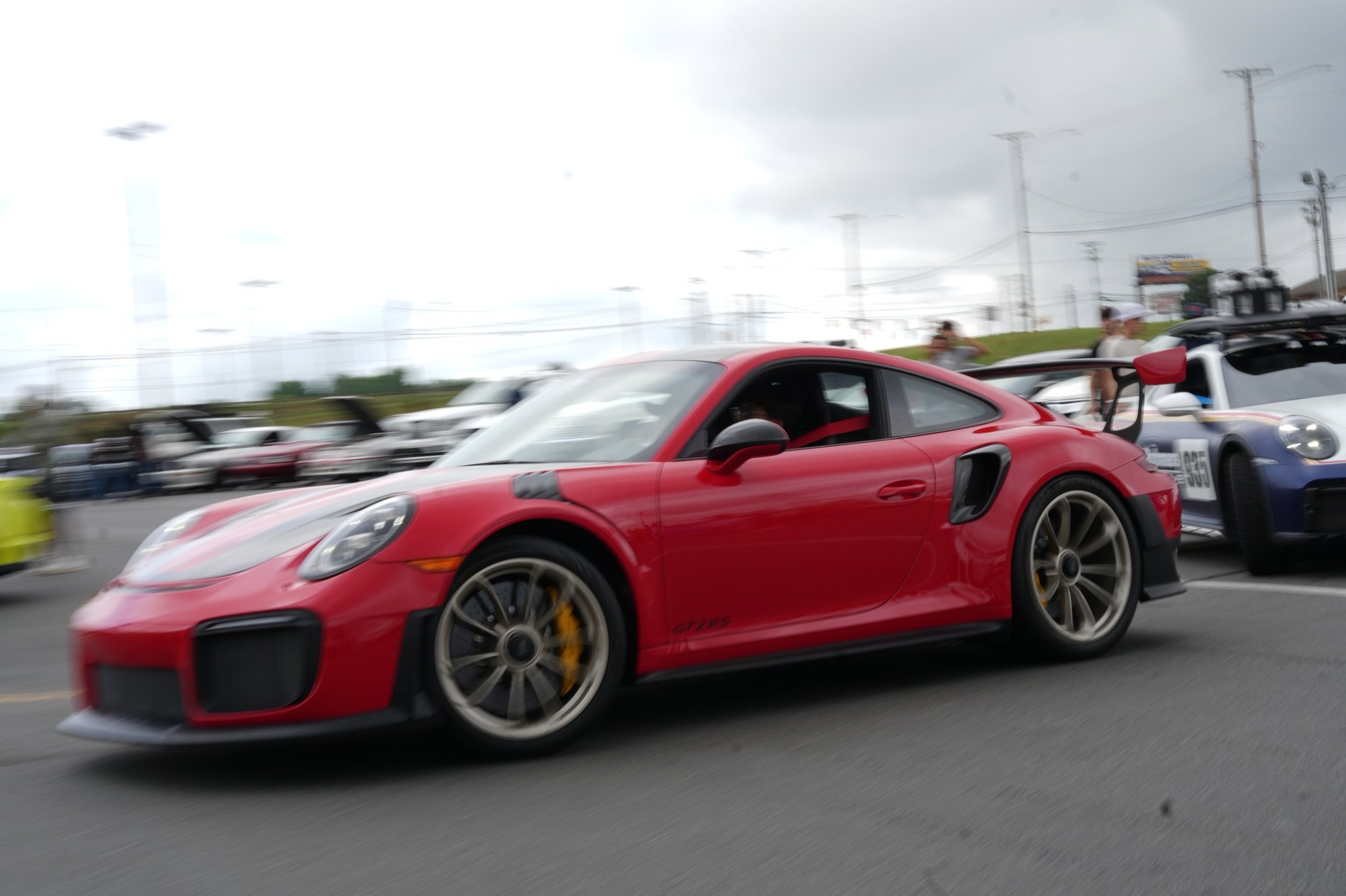 Red Porsche 911 GT3 replica car with black accents and large rear wing on a racetrack or car event with other sports cars in the background