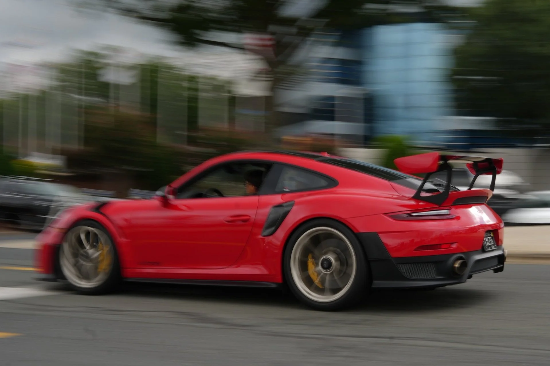 Red sports car with a large rear wing in motion on a city street, background trees and buildings blurred.