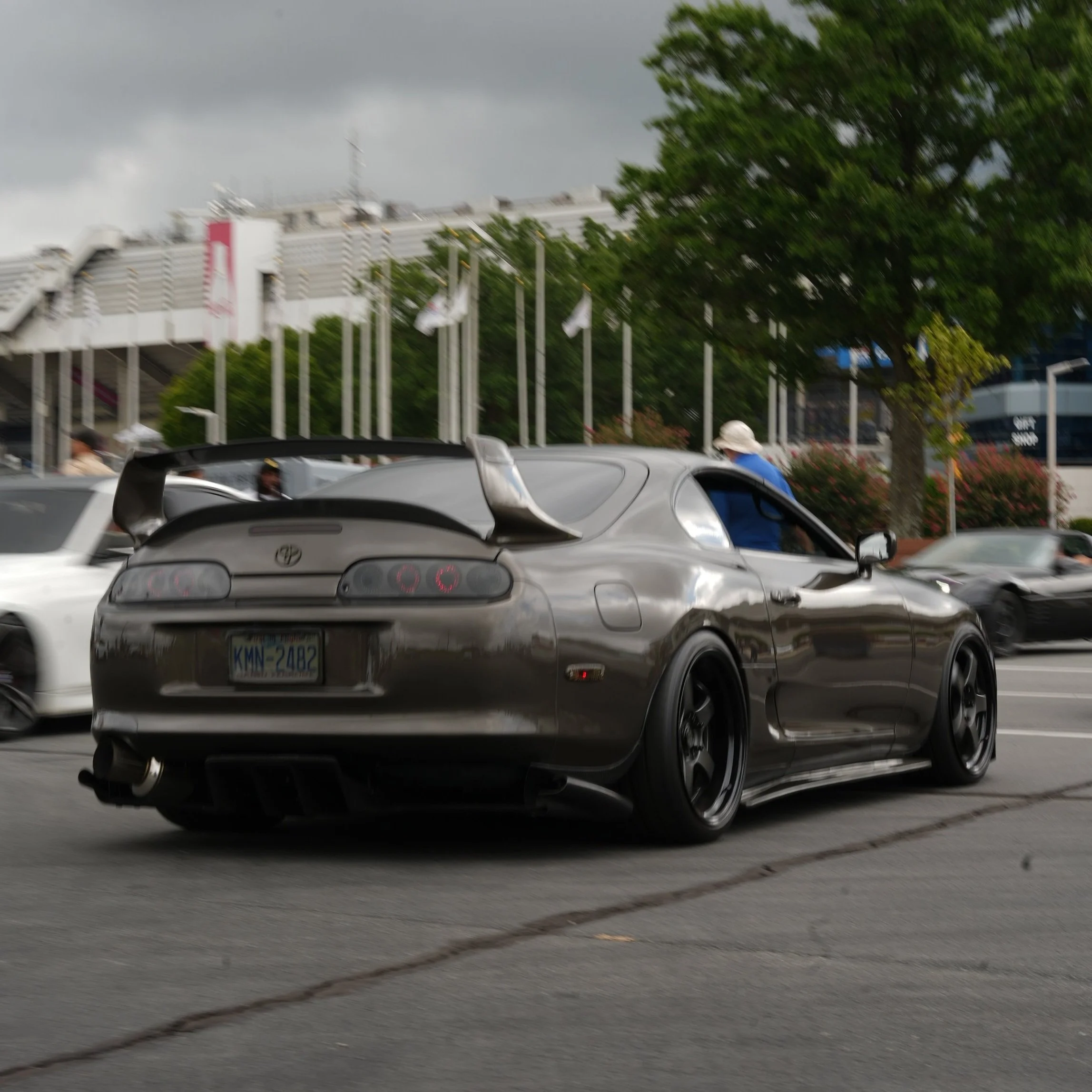 A gray Toyota Supra sports car with a large rear spoiler, black rims, and custom side skirts on a city street, with other cars and trees in the background.
