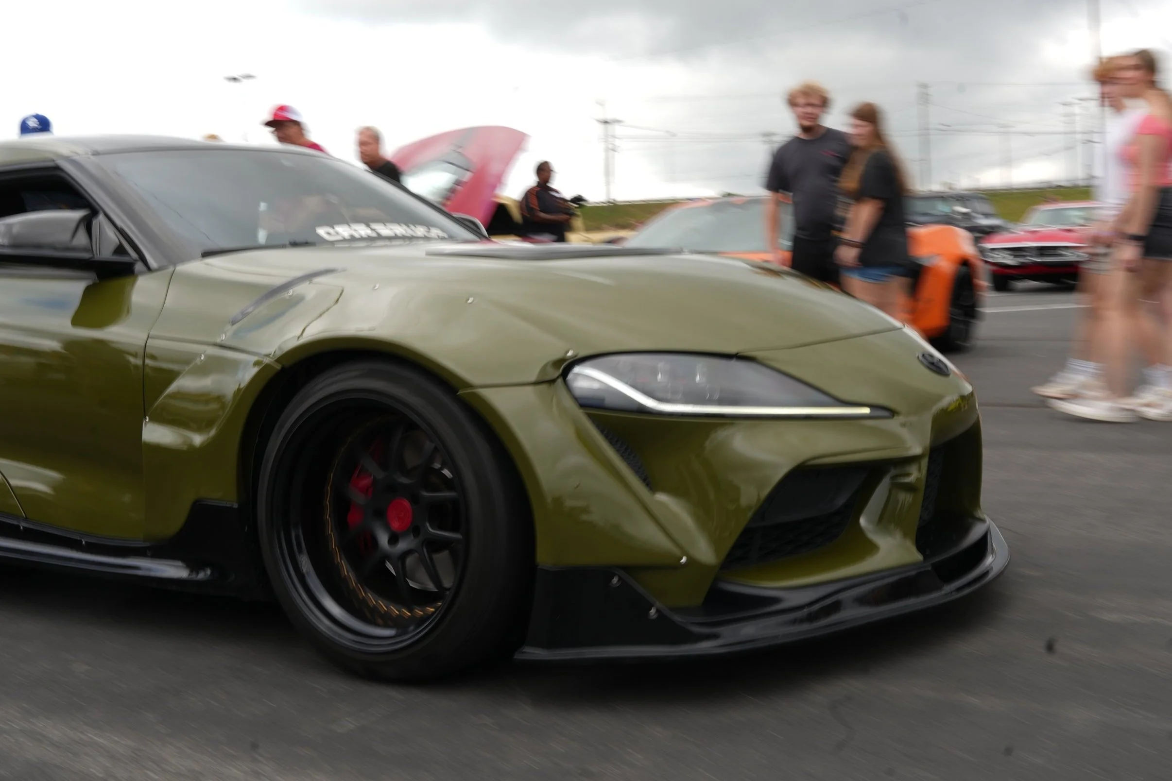 A green sports car on display at a car show, with several people looking at it in the background, some blurred due to motion or focus.