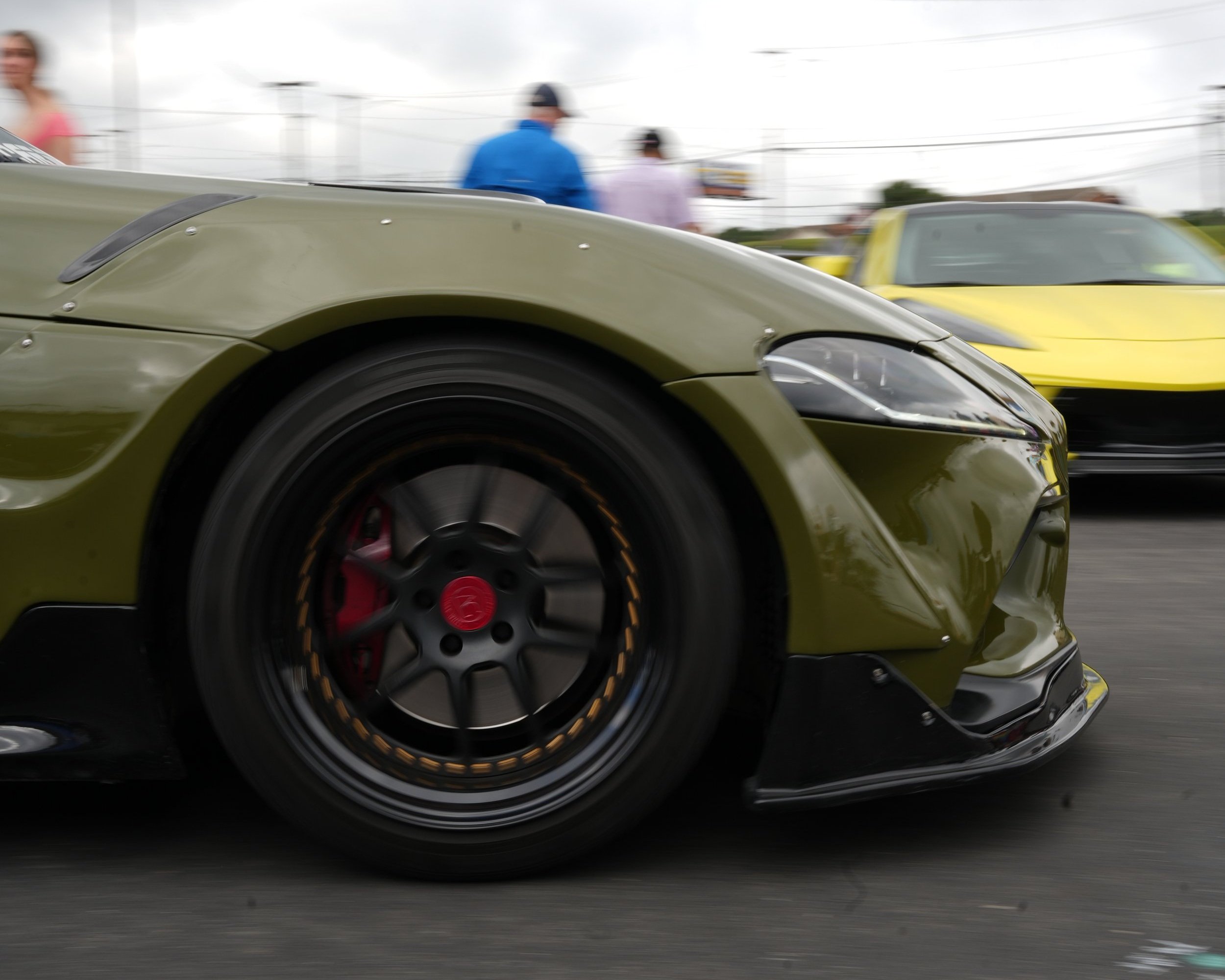 Close-up of a green racing car's front wheel and body, with a yellow car and people in the background at a racing event.