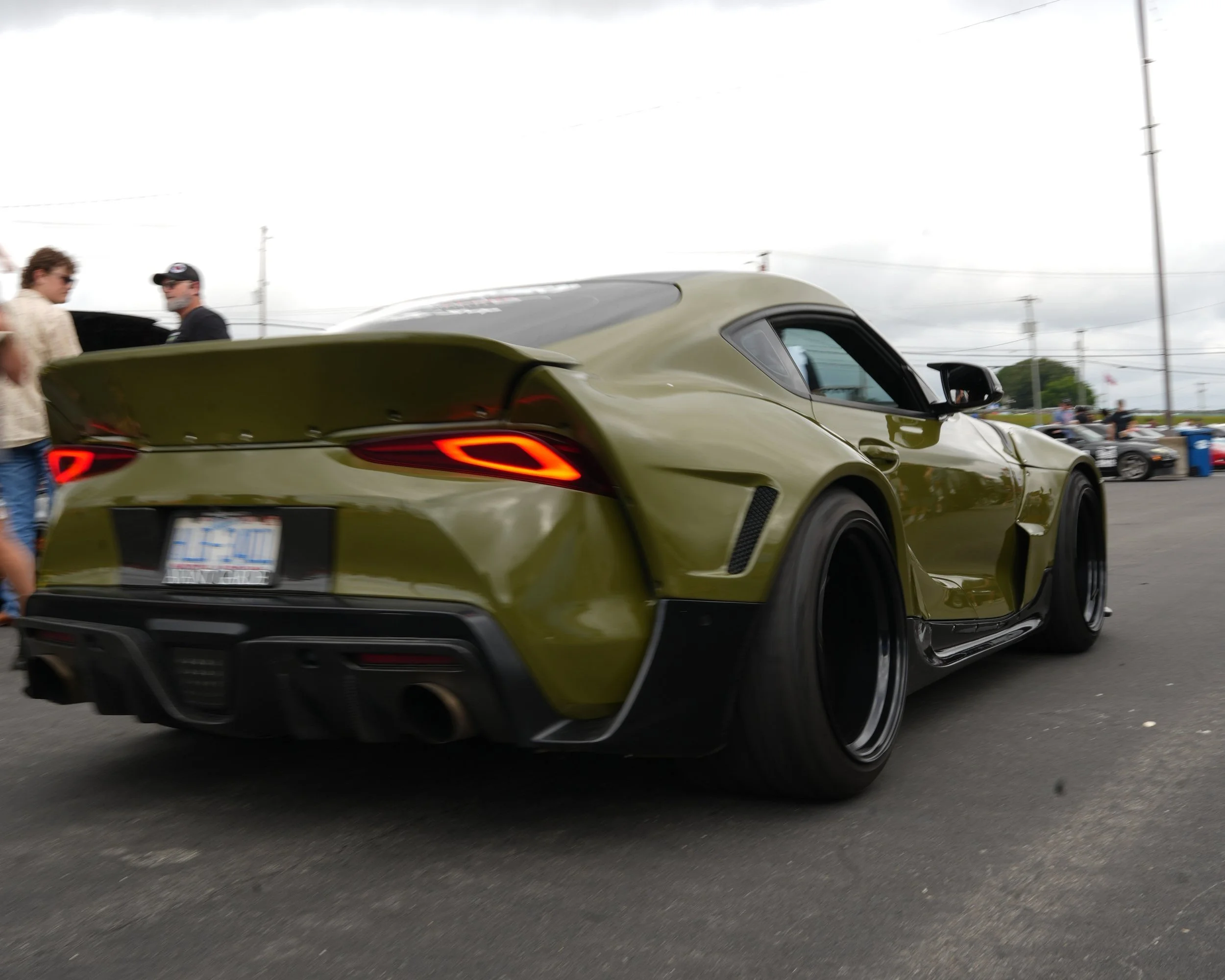 Green sports car with wide black tires at a car meet, with people and other vehicles in the background.