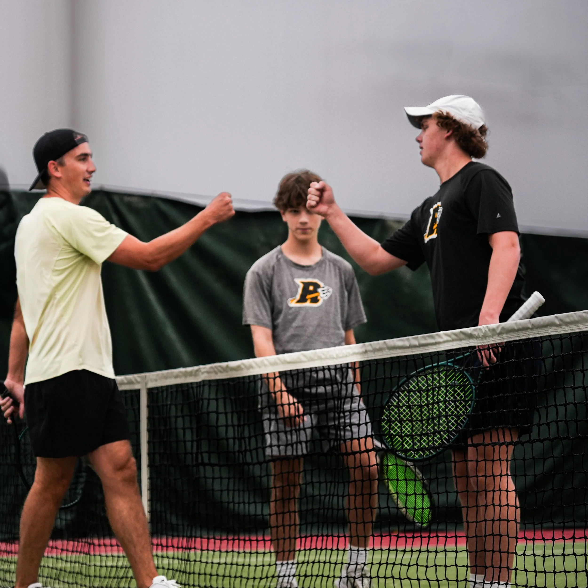 Two young men shaking hands after a tennis match on an indoor court, with a younger boy standing between them observing.