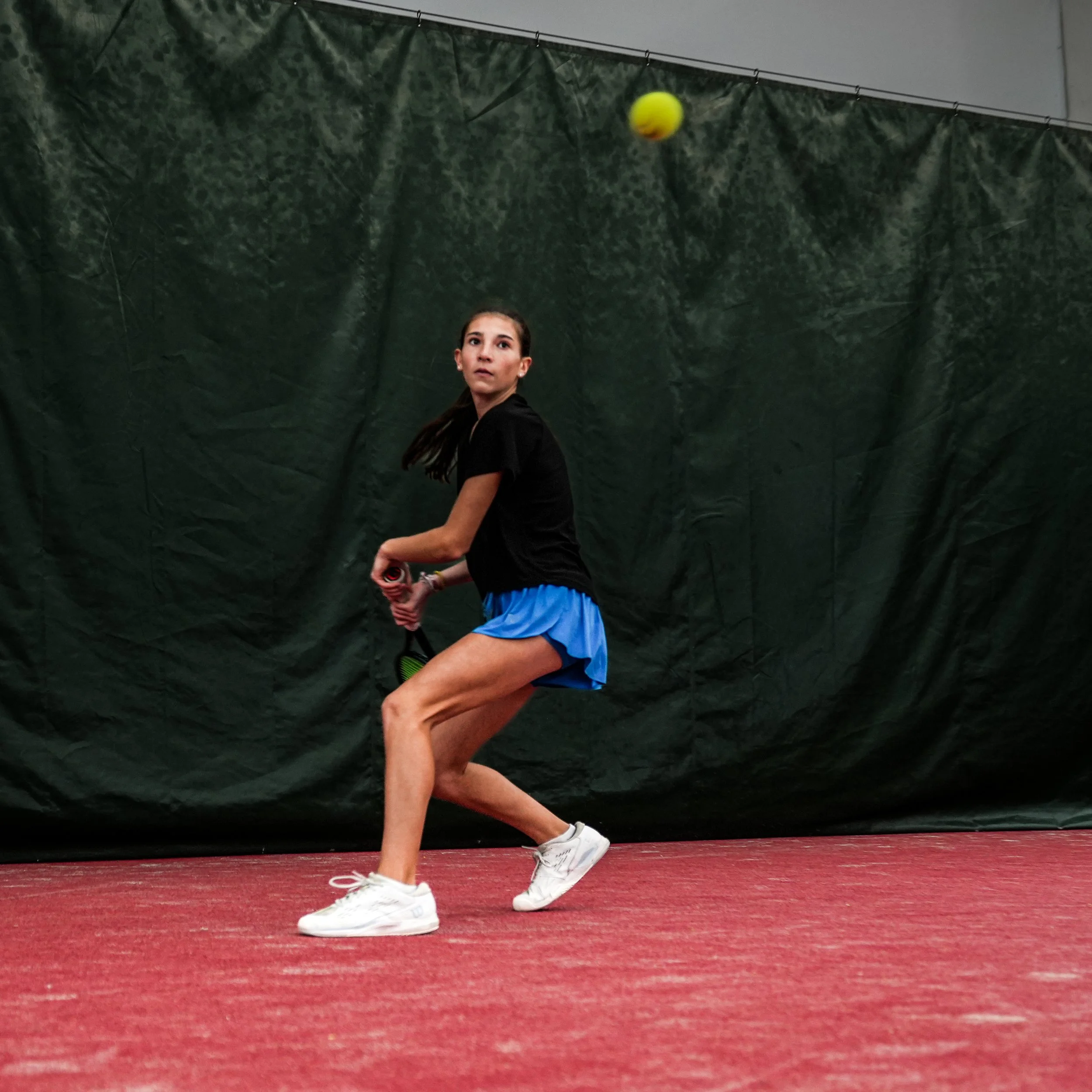 A young female tennis player is preparing to hit a yellow tennis ball inside an indoor tennis court with a dark green backdrop and red flooring.