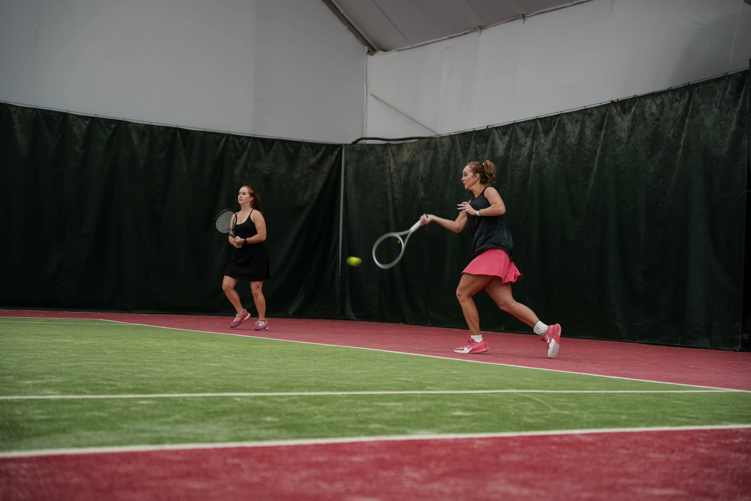 Two women playing tennis indoors on a court with green and red surfaces, one hitting a tennis ball with her racket, the other standing with her racket, near a black curtain background.