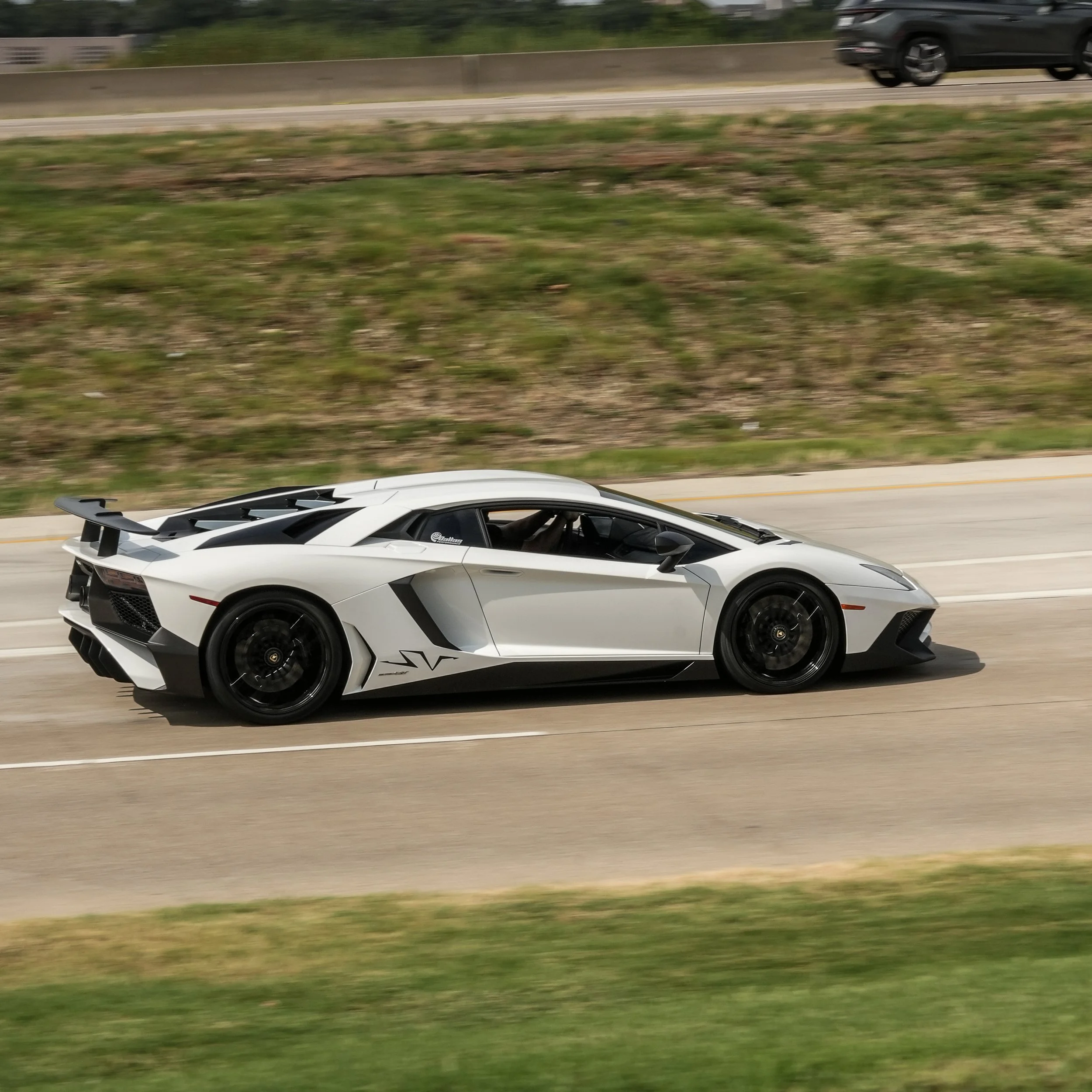 White Lamborghini sports car on a highway with a black rear spoiler and black wheels, blurred background indicating motion.