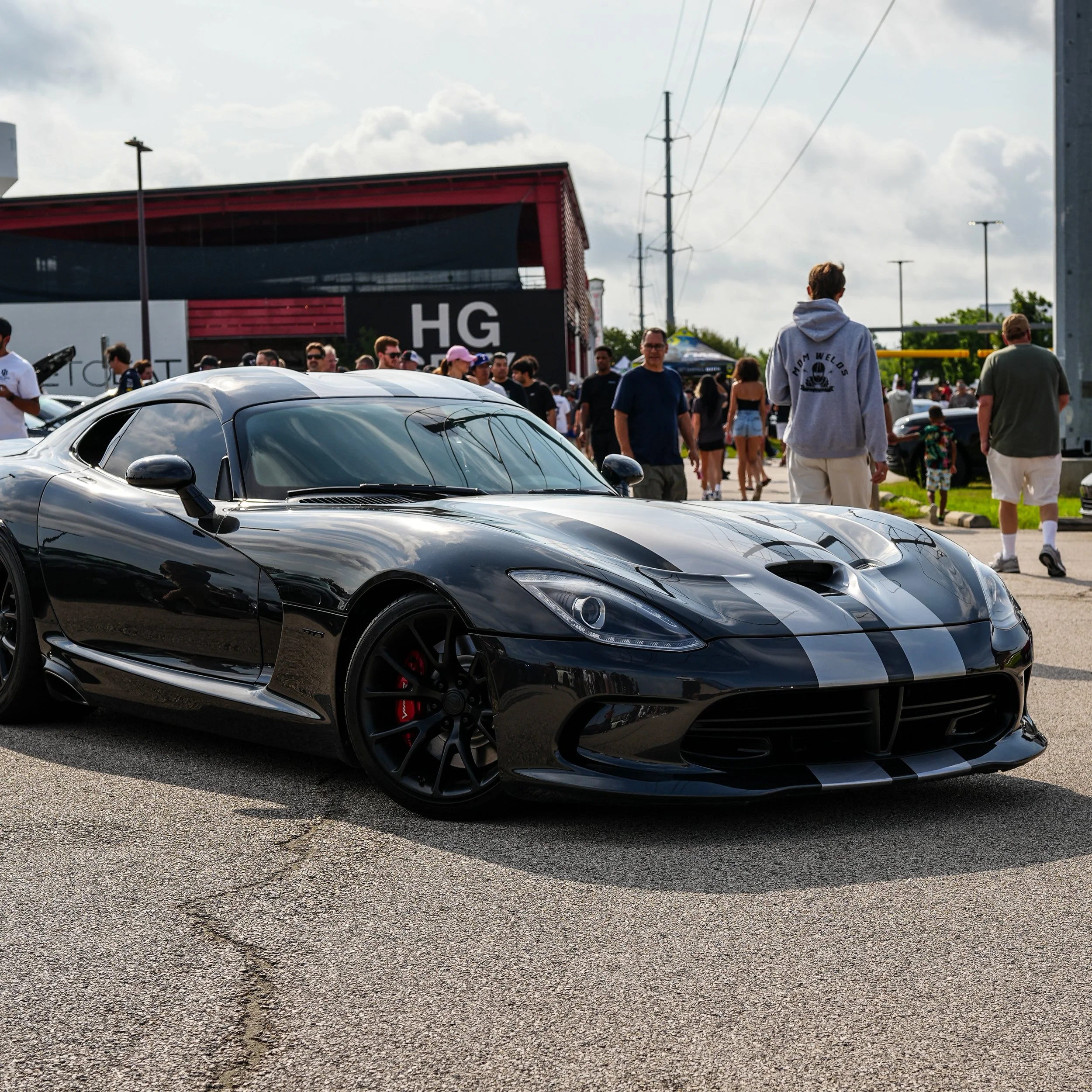 A black sports car with racing stripes parked at an outdoor car show, with people walking and observing vehicles in the background.