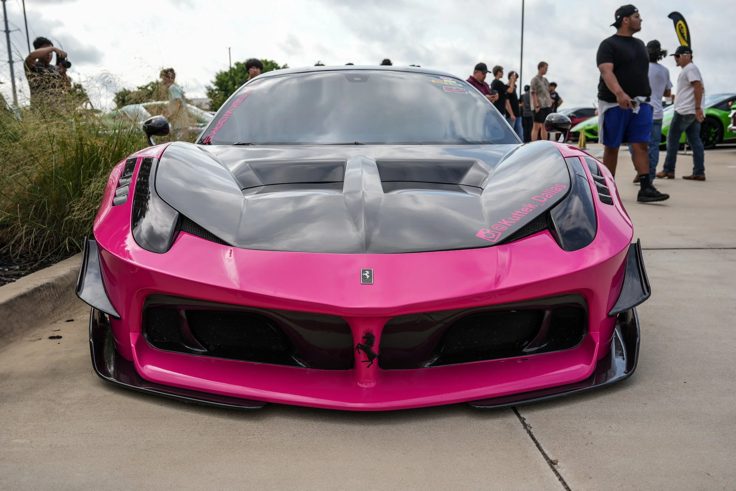 Pink Ferrari sports car with black carbon fiber hood on display at a car show, with people standing in the background.