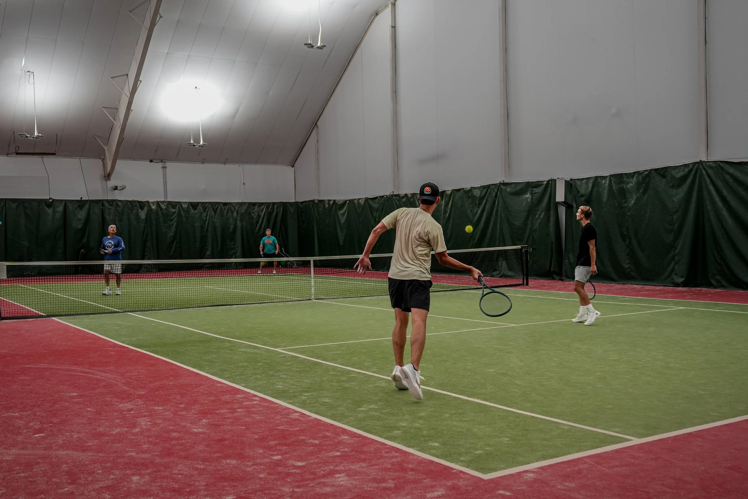 Four men playing indoor tennis on a green court with a net in the middle. The court has a red border, and the background features dark green curtains and ceiling lights.