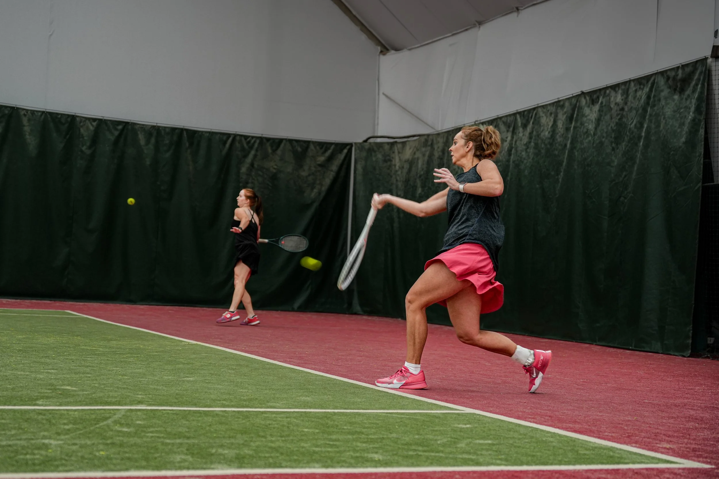 Two women playing tennis indoors on a red and green court, practicing forehand shots.