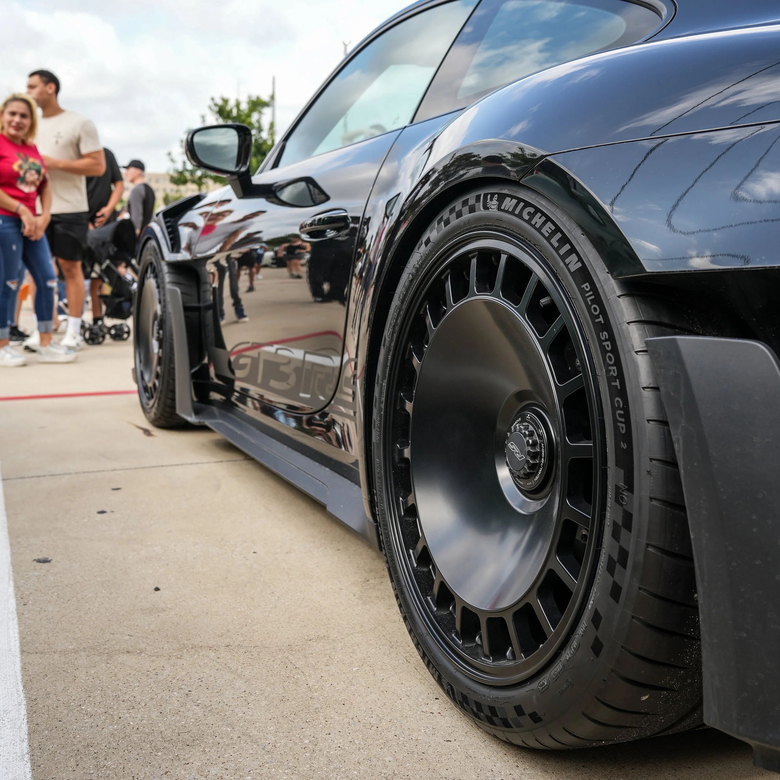 Close-up of a black race car with racing tires, parked outdoors with people in the background.