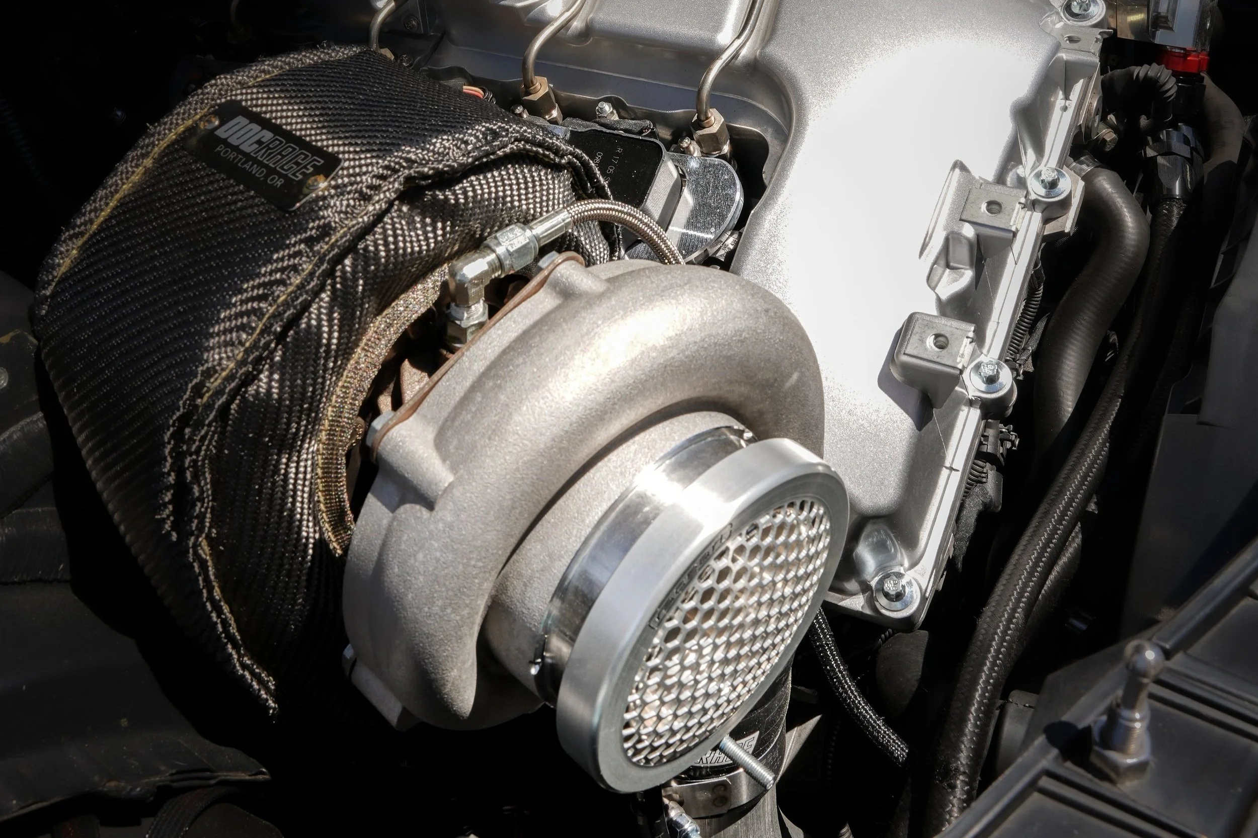 Close-up view of a turbocharger installed in a car engine with silver components, black hoses, and a heat shield marked with DOC Race Portland.