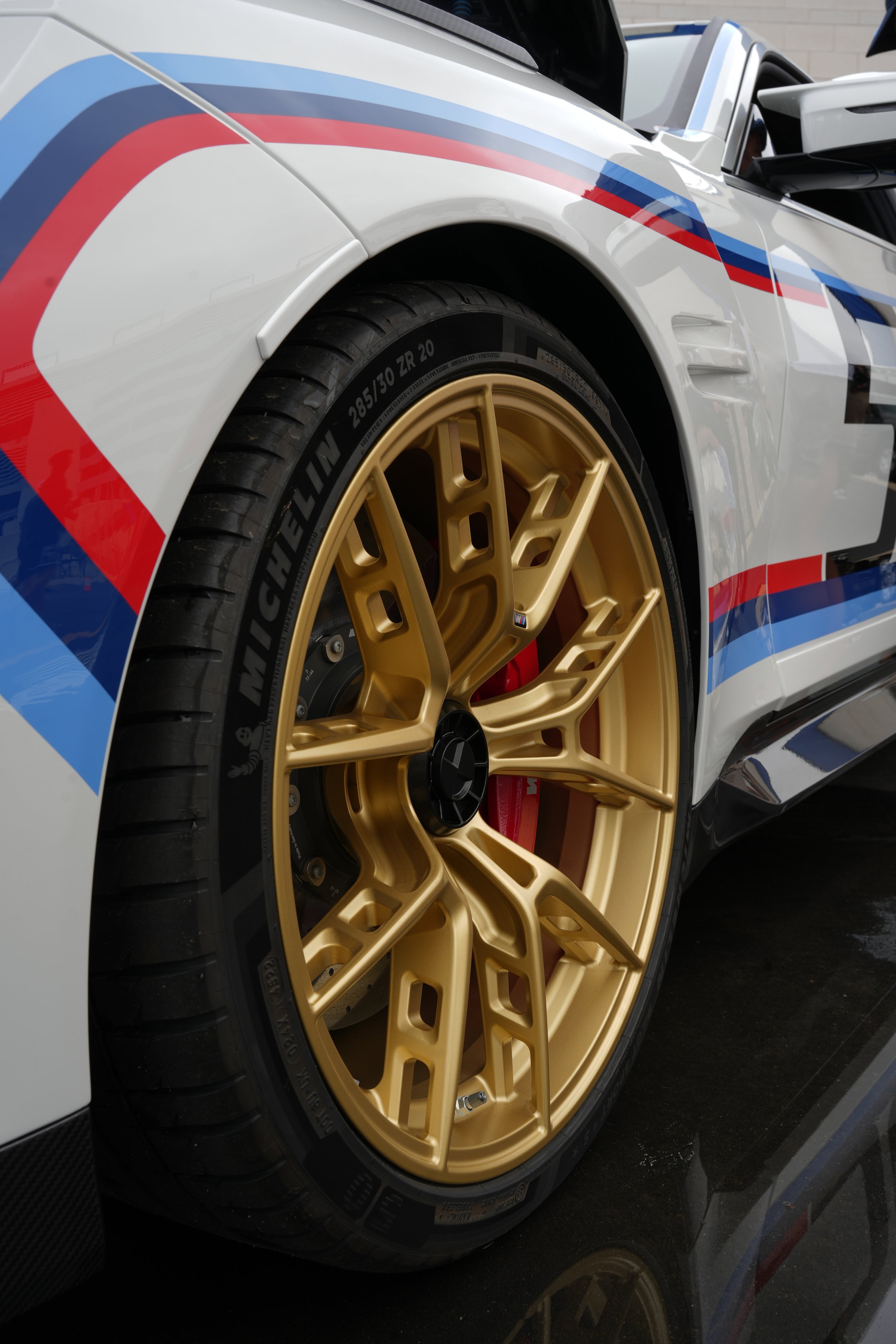 Close-up of a luxury sports car wheel with a gold rim, tire, and red brake caliper, part of a race car with racing stripes in red, blue, and white.