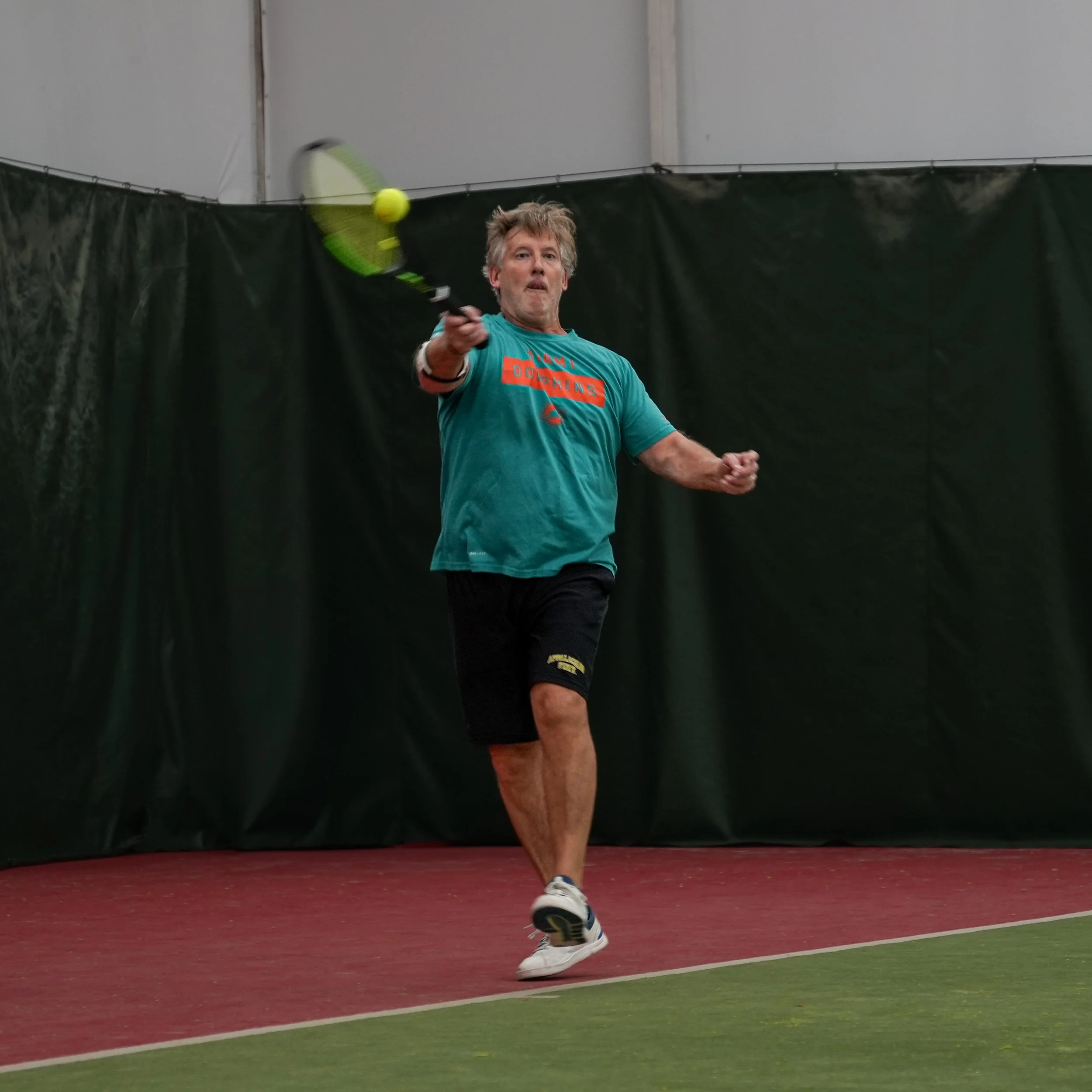 Man playing tennis indoors, preparing to hit a tennis ball with a racket, on a court with green and red flooring.