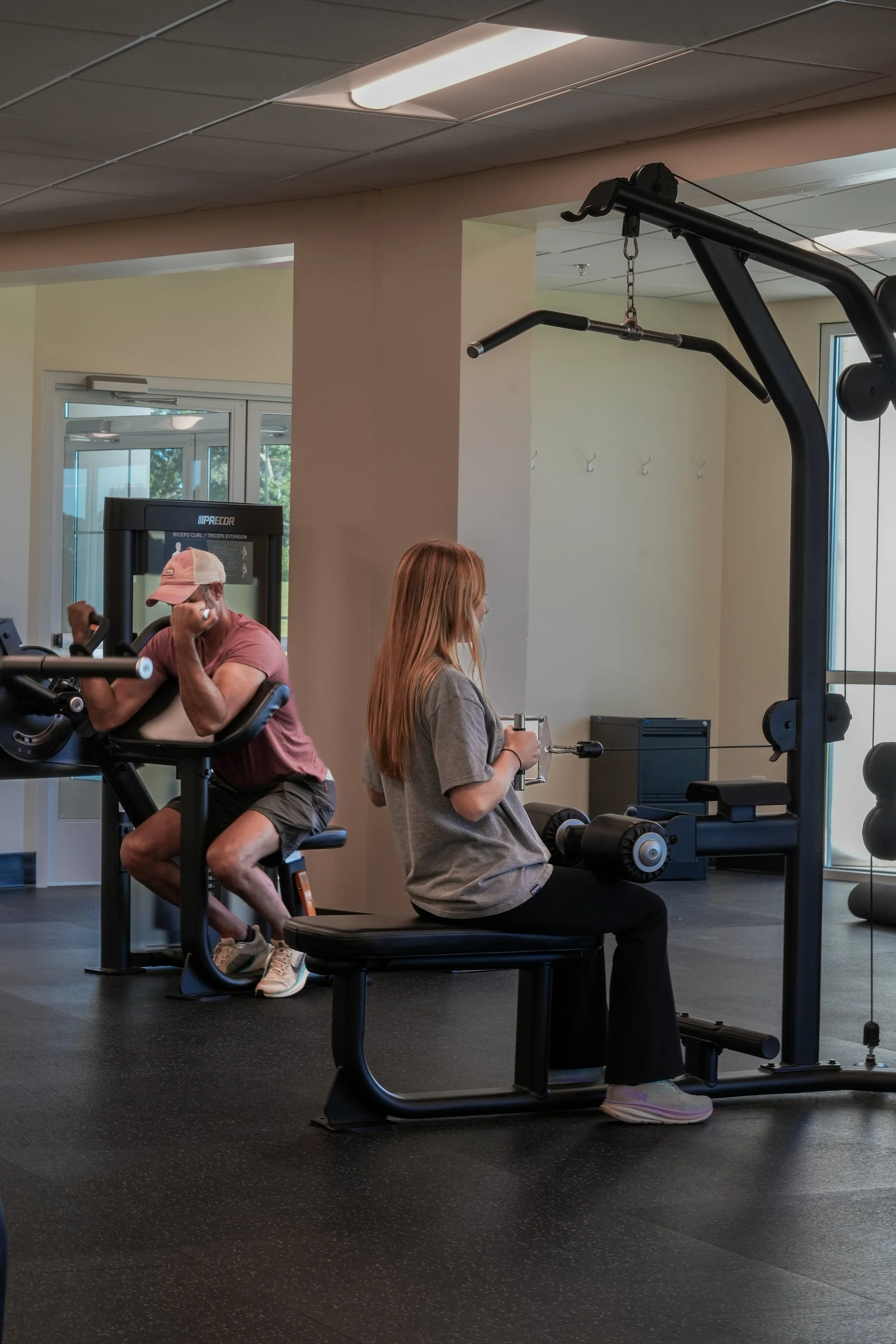 A woman using a seated chest press machine while a man on a nearby exercise machine in a gym.