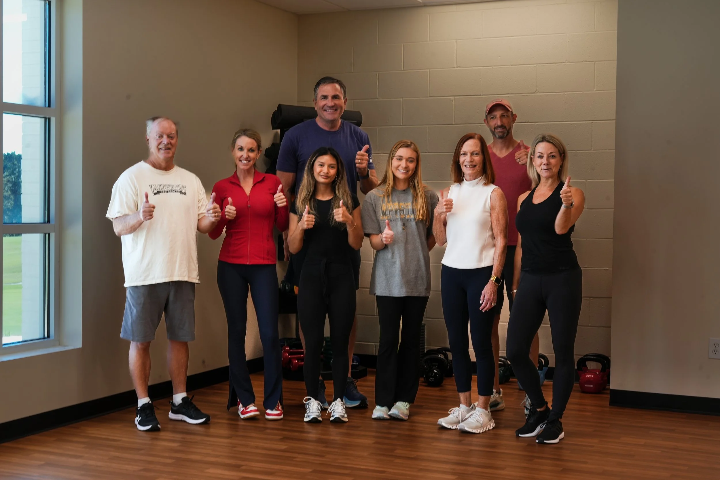 Group of nine people in workout attire posing indoors, giving thumbs-up.