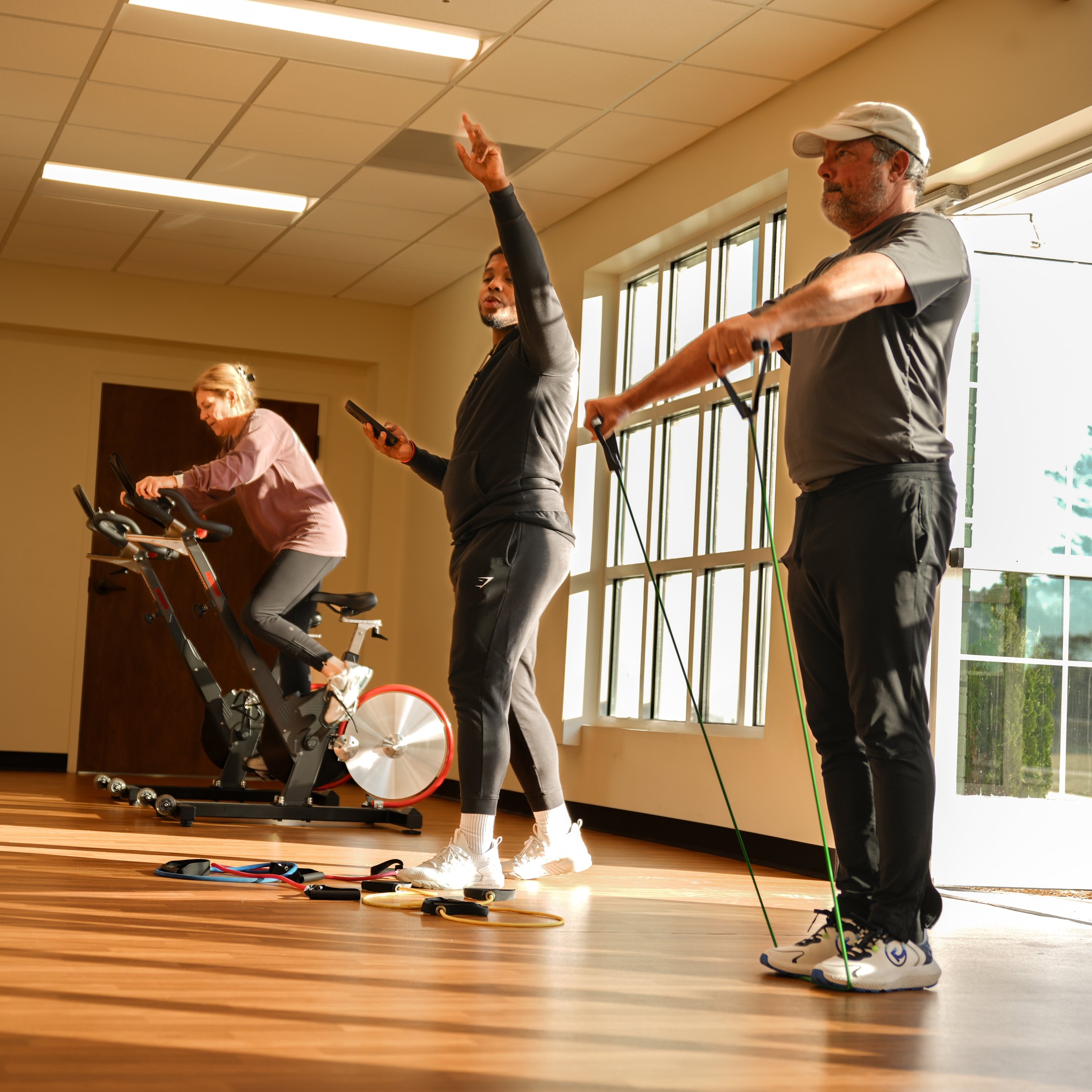 Three people exercising indoors: a woman on a stationary bike, a man holding a smartphone and standing, and another man using resistance bands, in a brightly lit room with large windows.