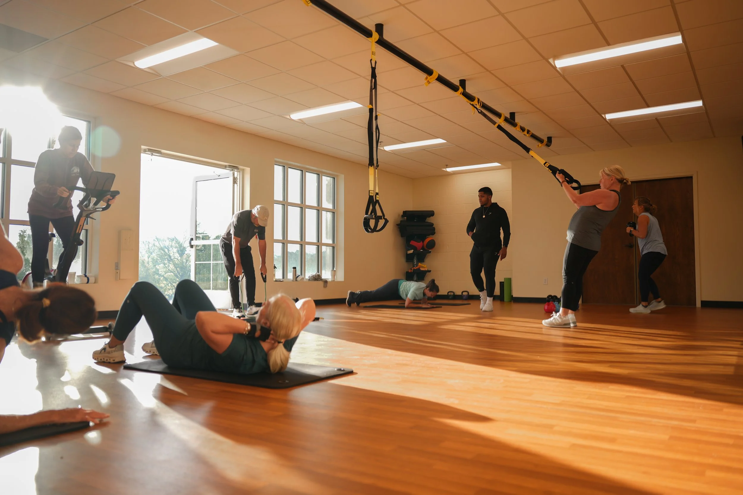 Group of people participating in a fitness class indoors. Some are exercising on mats, while others are using gym equipment and TRX straps hanging from the ceiling. Bright natural light coming through large windows.
