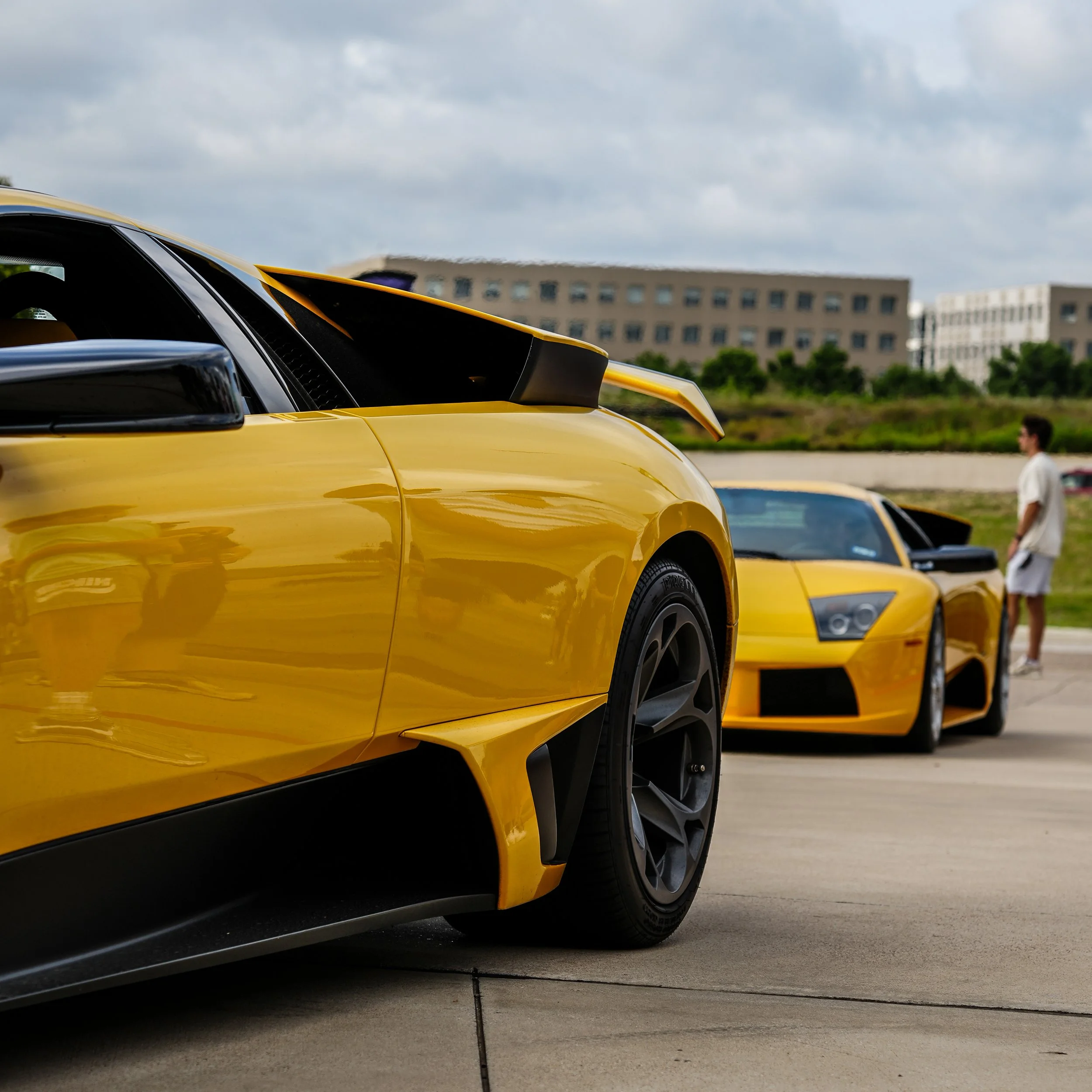 Close-up of a yellow luxury sports car, with another similar yellow sports car in the background, parked outdoors with a person walking nearby.