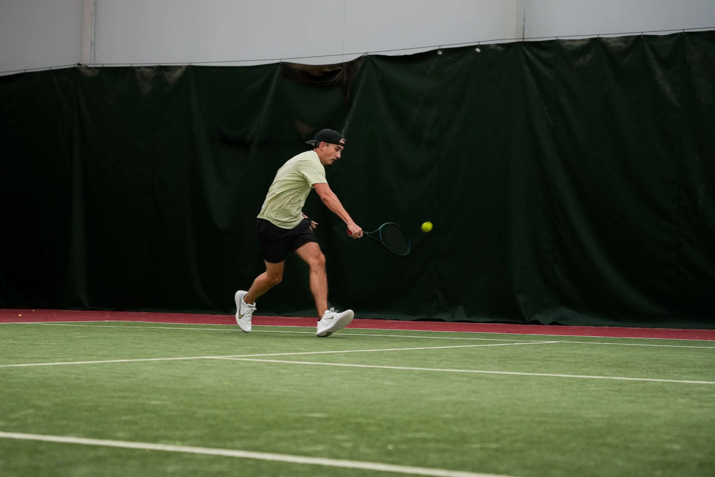 Man playing indoor tennis, hitting a yellow tennis ball with a racket on a green court.