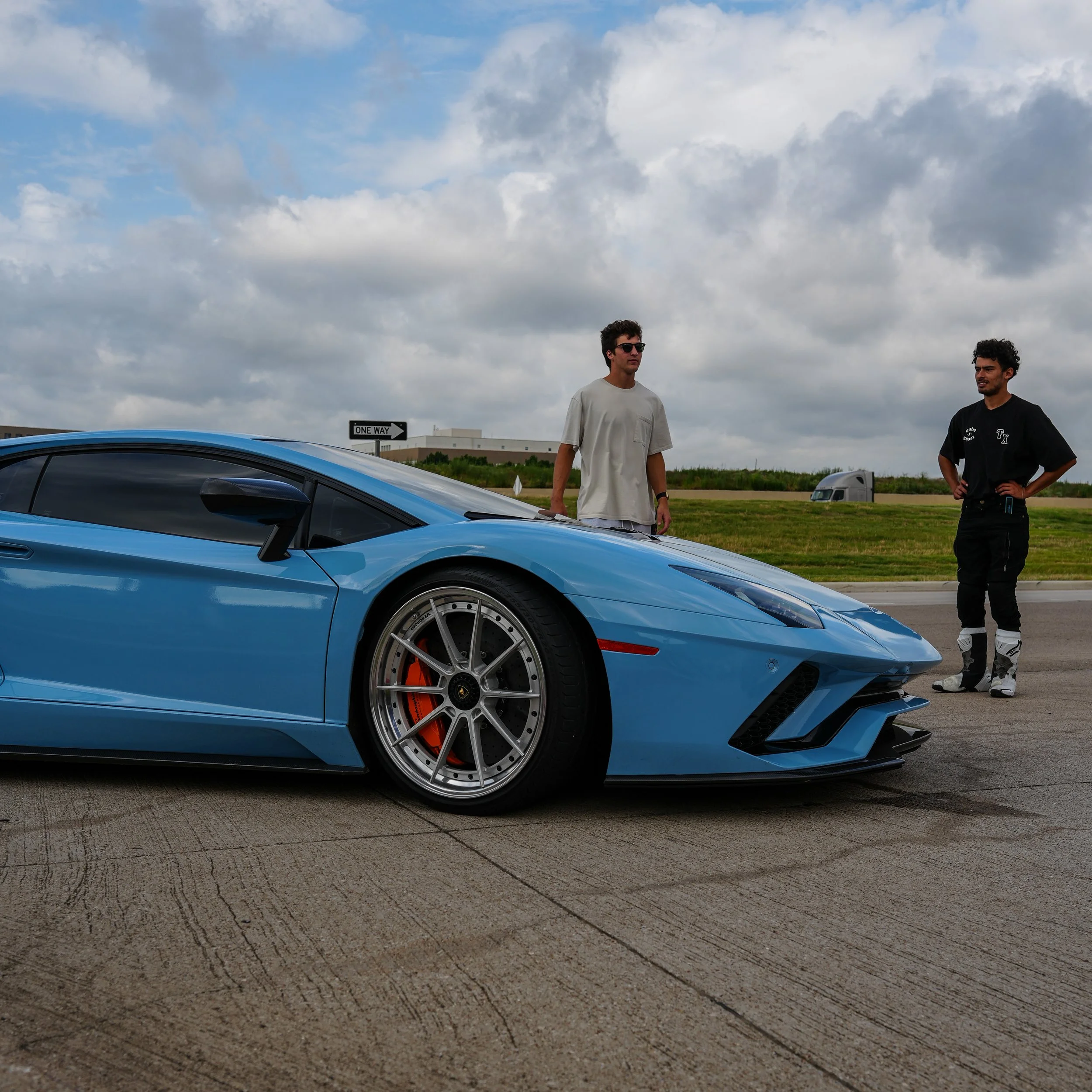 A blue Lamborghini sports car parked on a concrete surface with two men standing nearby, one in a white t-shirt and sunglasses, and the other in racing gear. The background features a grassy area, a sign saying 'One Way,' and a partly cloudy sky.