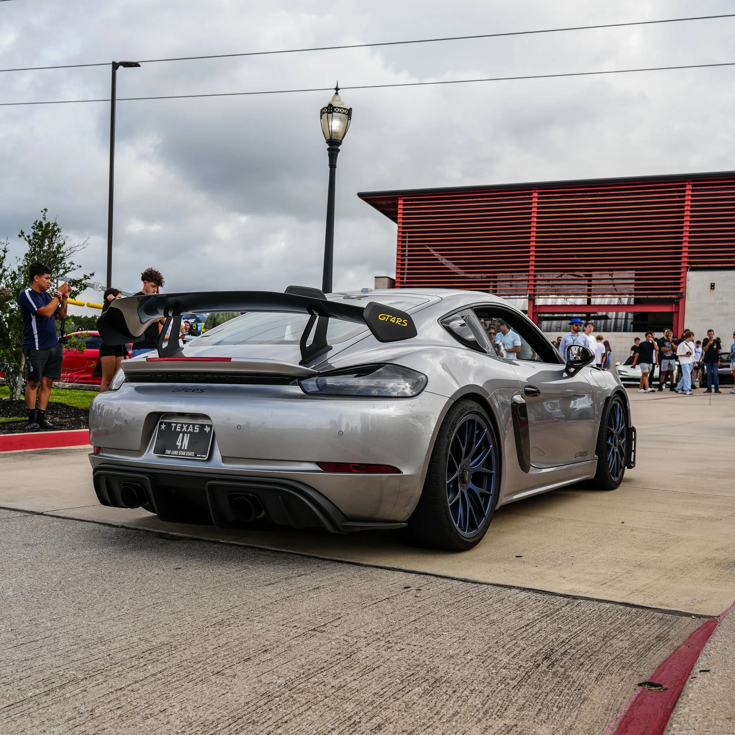 Silver sports car with large rear wing and black accent wheels parked on a concrete surface at an outdoor event with people in the background.