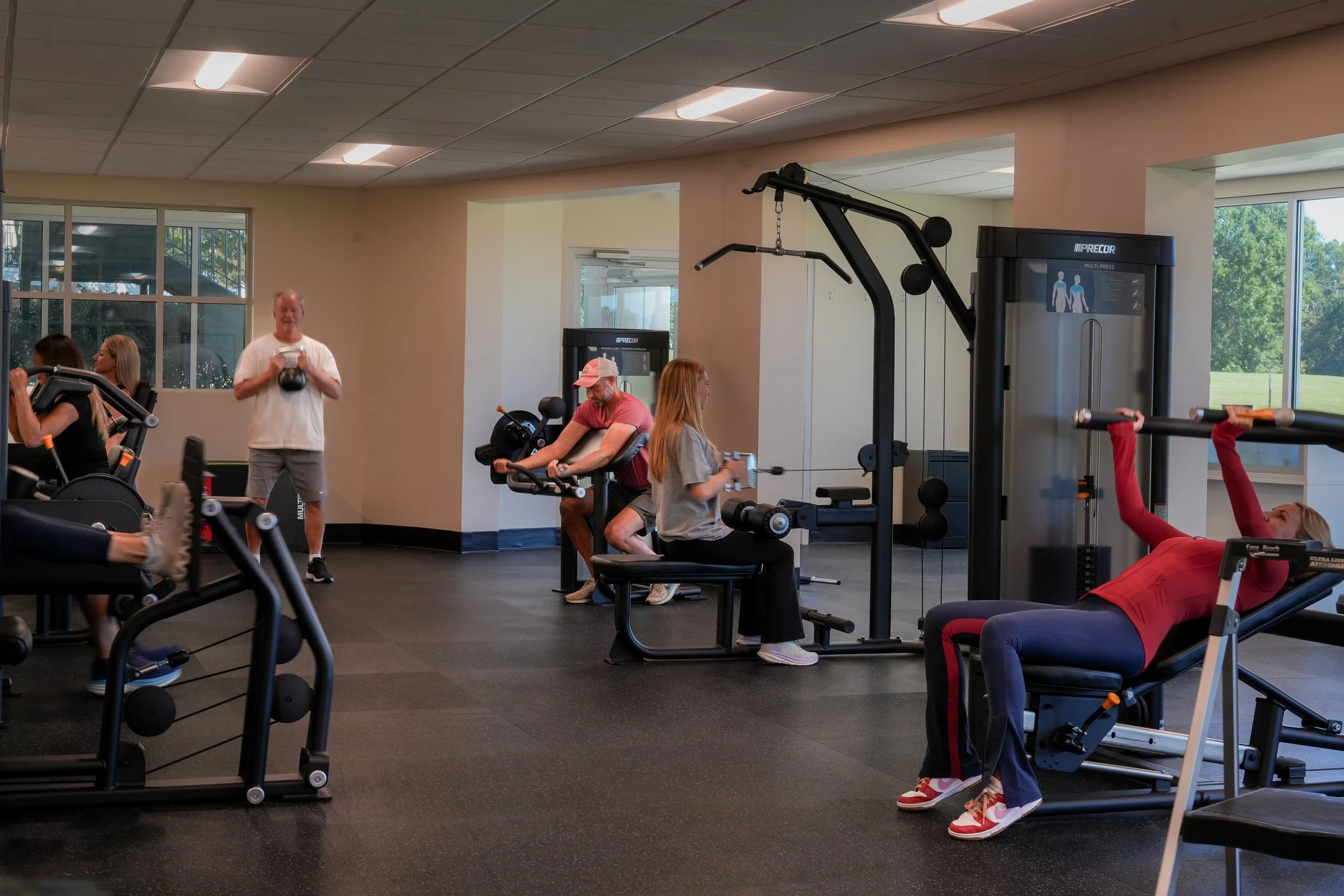 People working out with various exercise equipment in a gym, including ellipticals, weight machines, and benches, with large windows showing a green outdoor landscape.