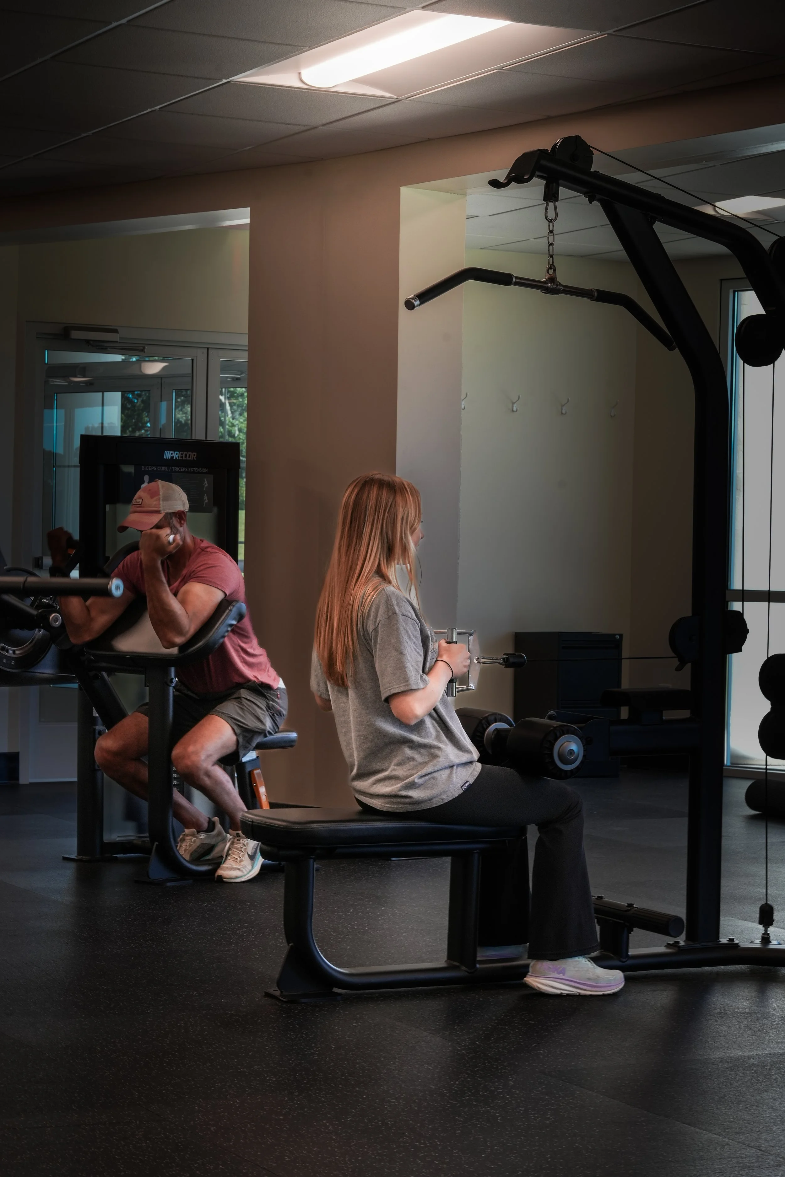 A young woman and a man working out at a gym, with the woman using a seated rowing machine and the man using a leg curl machine nearby.