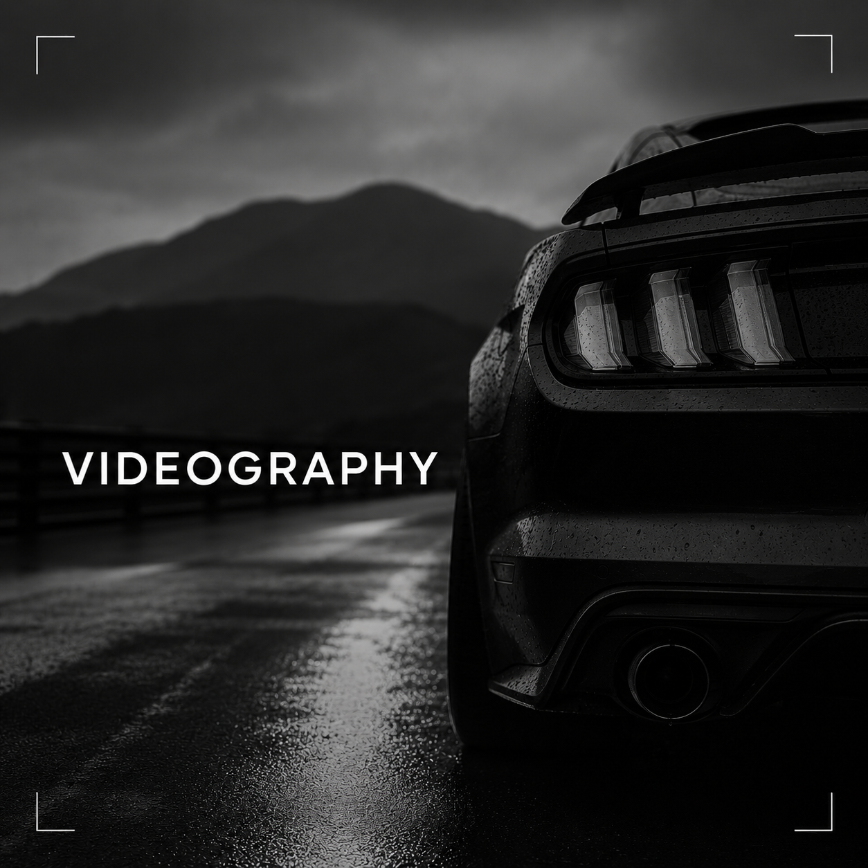 Close-up of the rear of a black sports car on a wet road with mountains in the background, rainy weather, and the word 'VIDEOGRAPHY' overlaid.