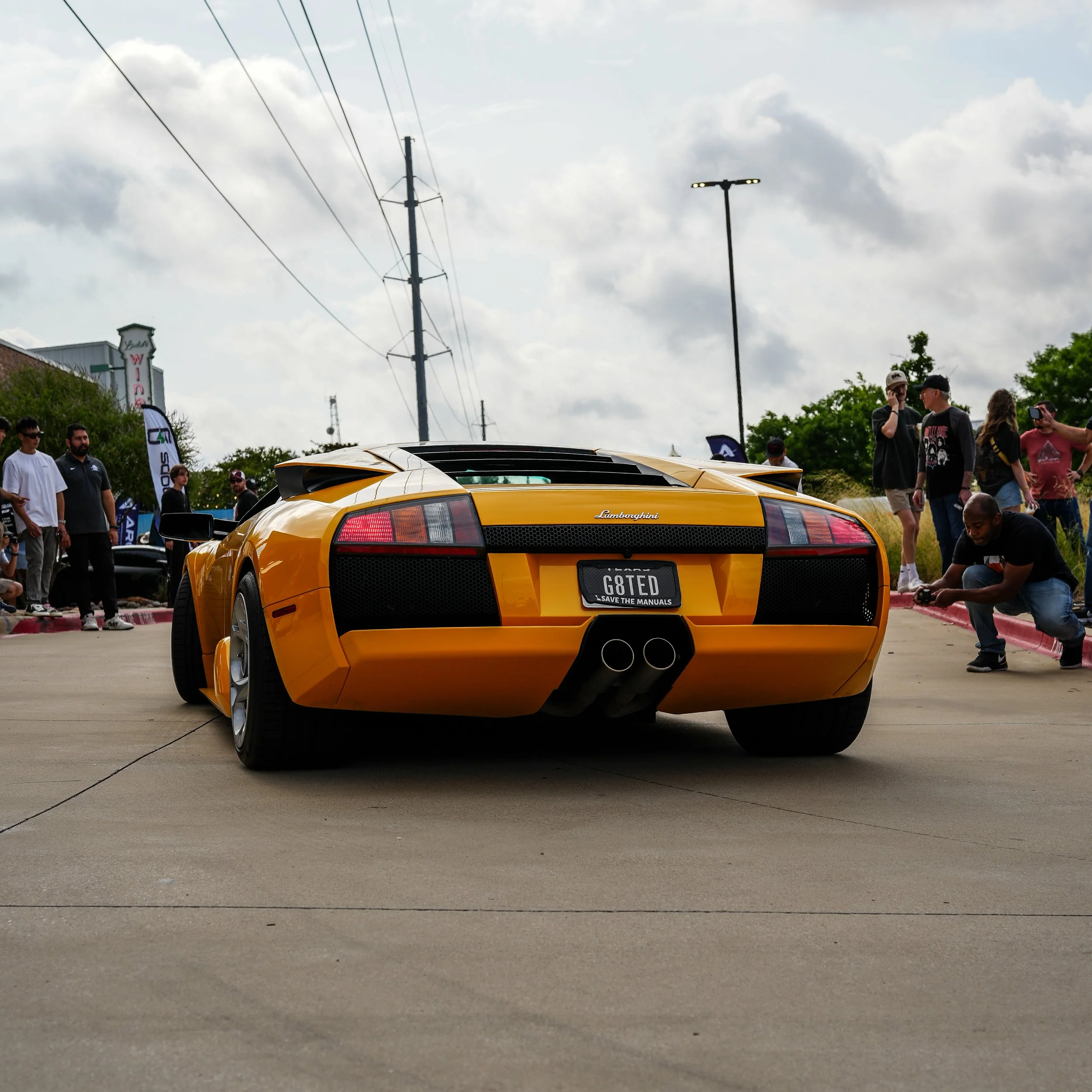A yellow Lamborghini sports car at a car show with people gathered around, some taking photos and videos.