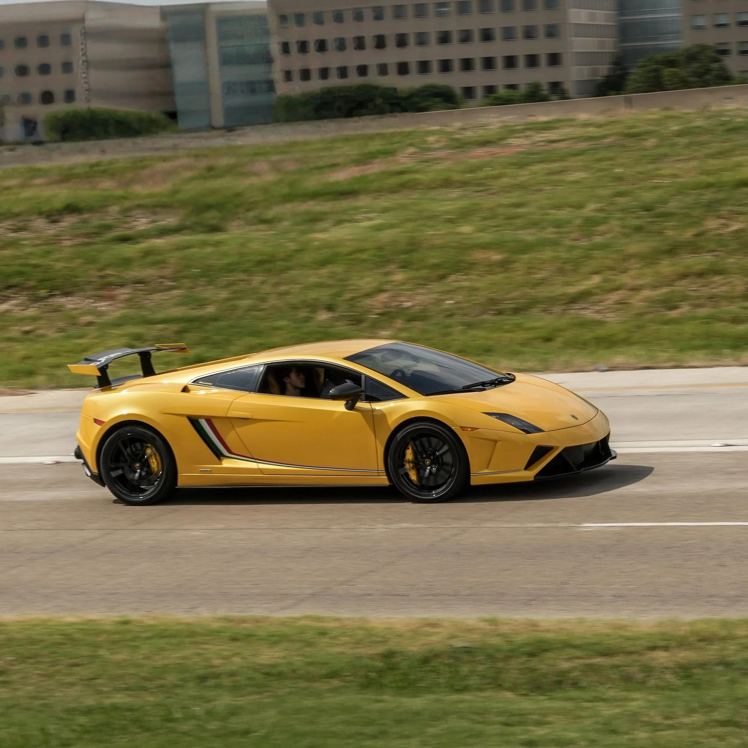 A yellow Lamborghini sports car driving on a city street with buildings and grass in the background.
