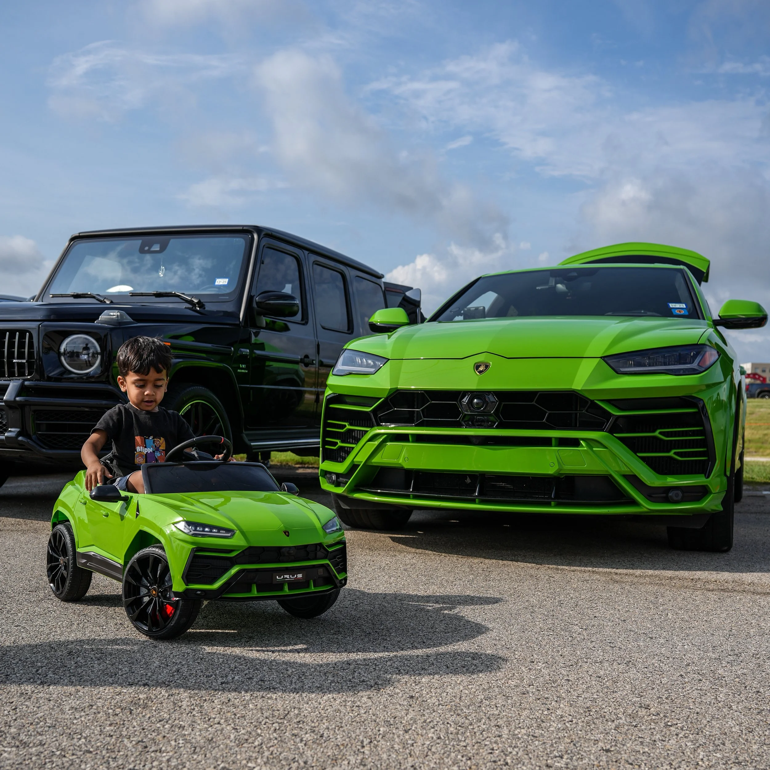A young boy sitting inside a green mini Lamborghini ride-on car, with a large green Lamborghini sports car and a black Mercedes-Benz G-Class vehicle parked behind him outdoors under a partly cloudy sky.