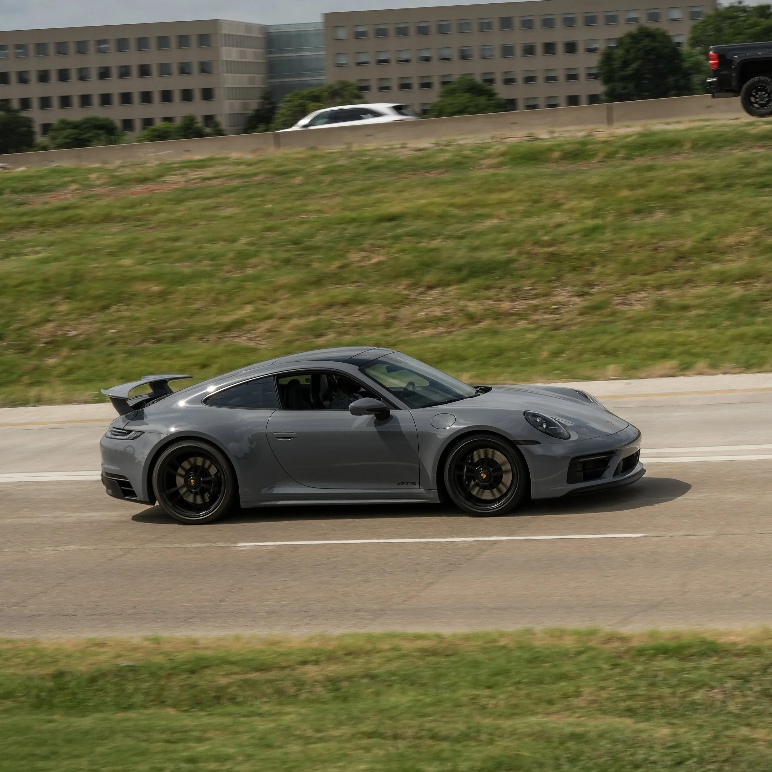 A gray sports car speeding on a multi-lane highway with a grassy hill and modern office buildings in the background.