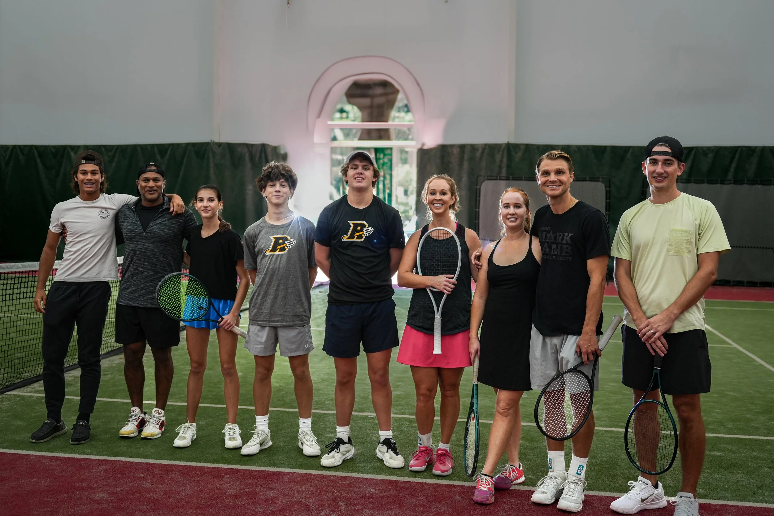 A group of ten young people standing together inside an indoor tennis court, holding tennis rackets and smiling at the camera.