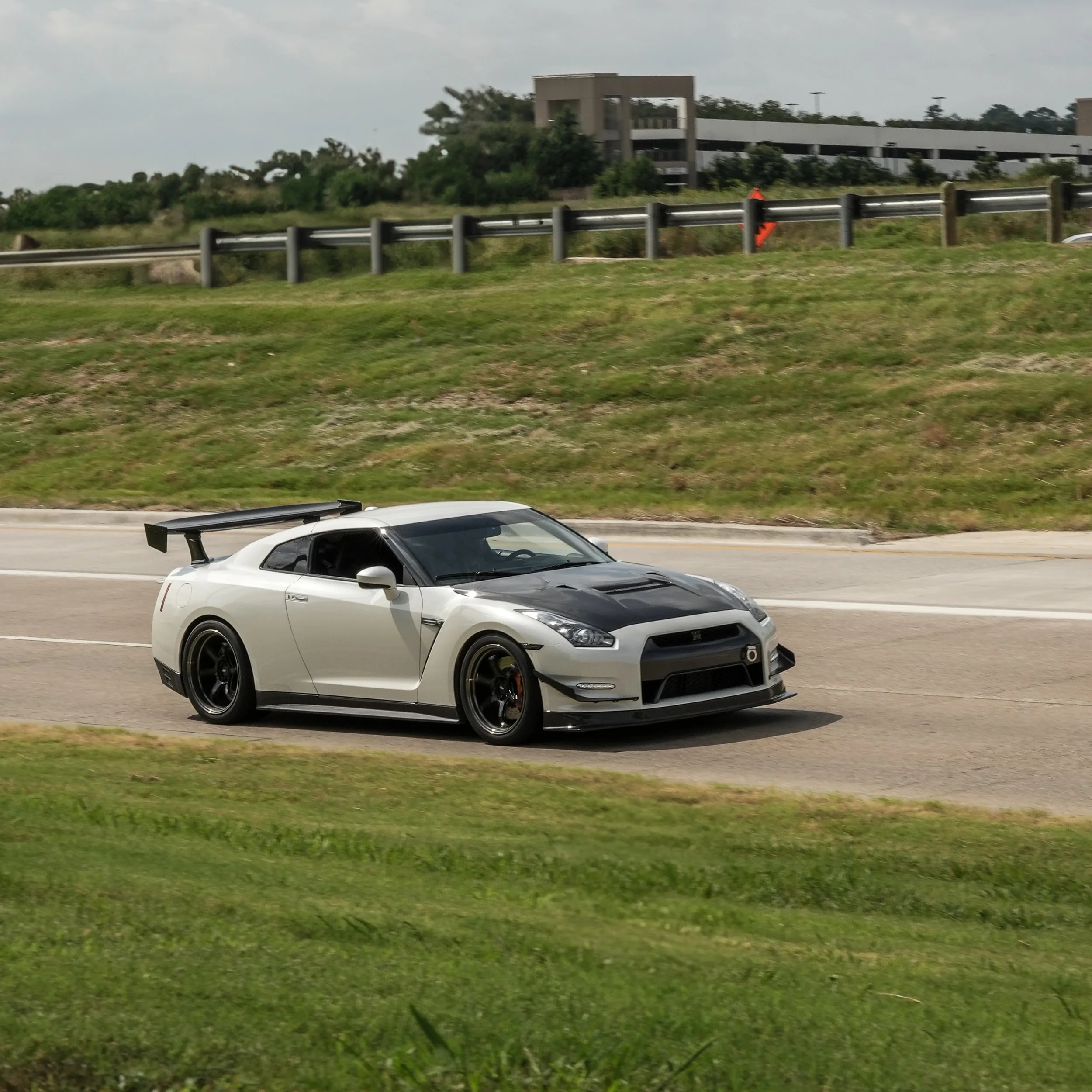A white and black sports car with a rear wing driving on a paved road in a grassy area.
