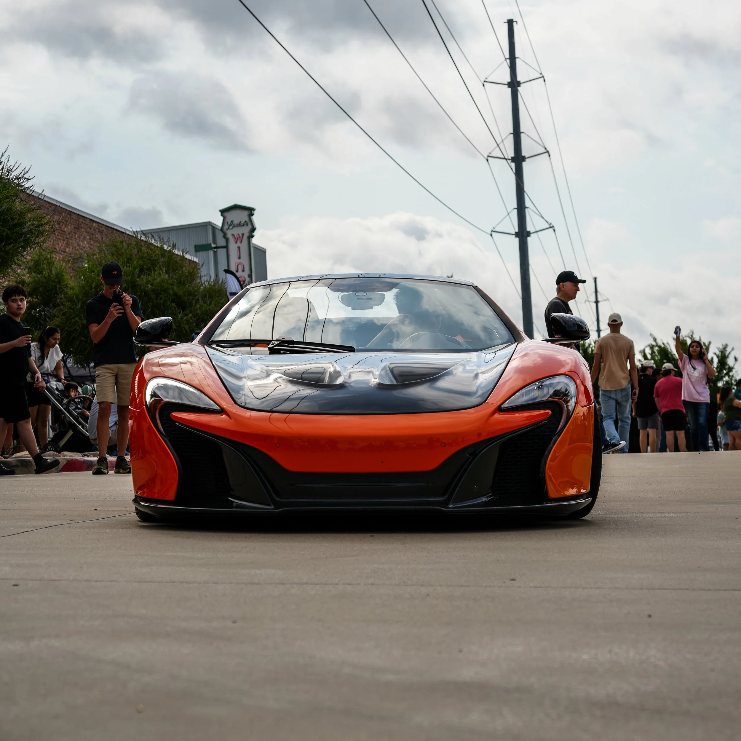 Orange sports car with black accents parked on concrete with people gathered around, some taking pictures, under cloudy sky.