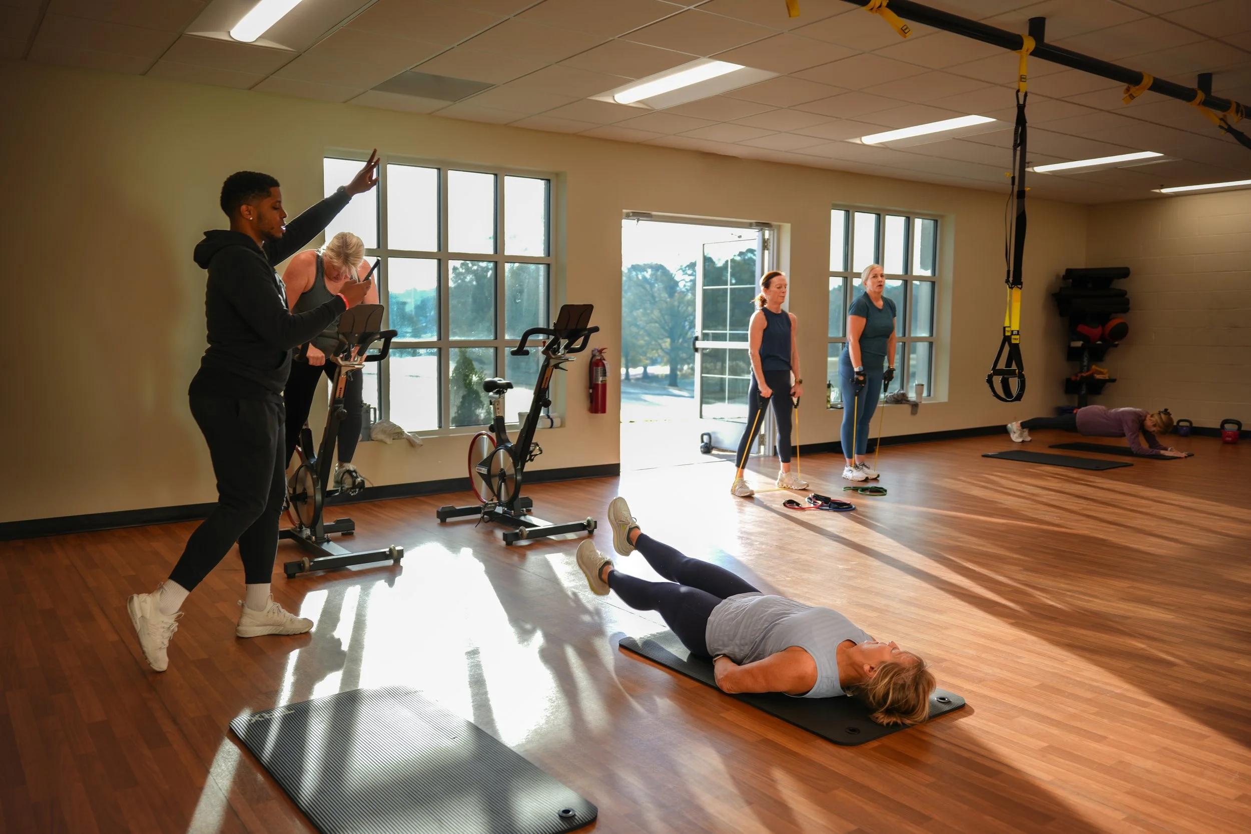 A group fitness class in a gym with parachute resistance training equipment, stationary bikes, and a woman doing a mat exercise while others observe or prepare for their workout.