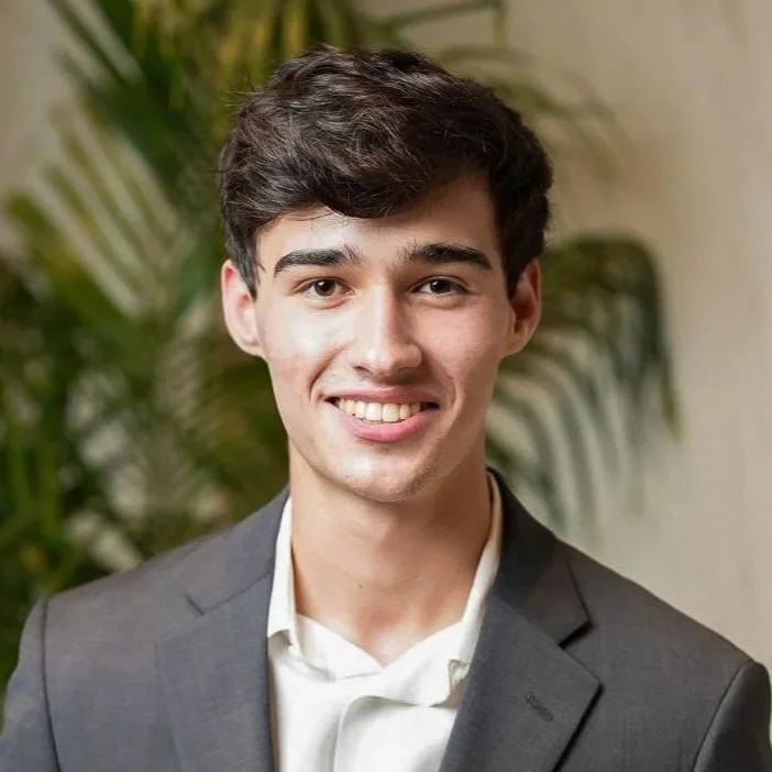 Young man smiling in a suit with a blurred plant background.