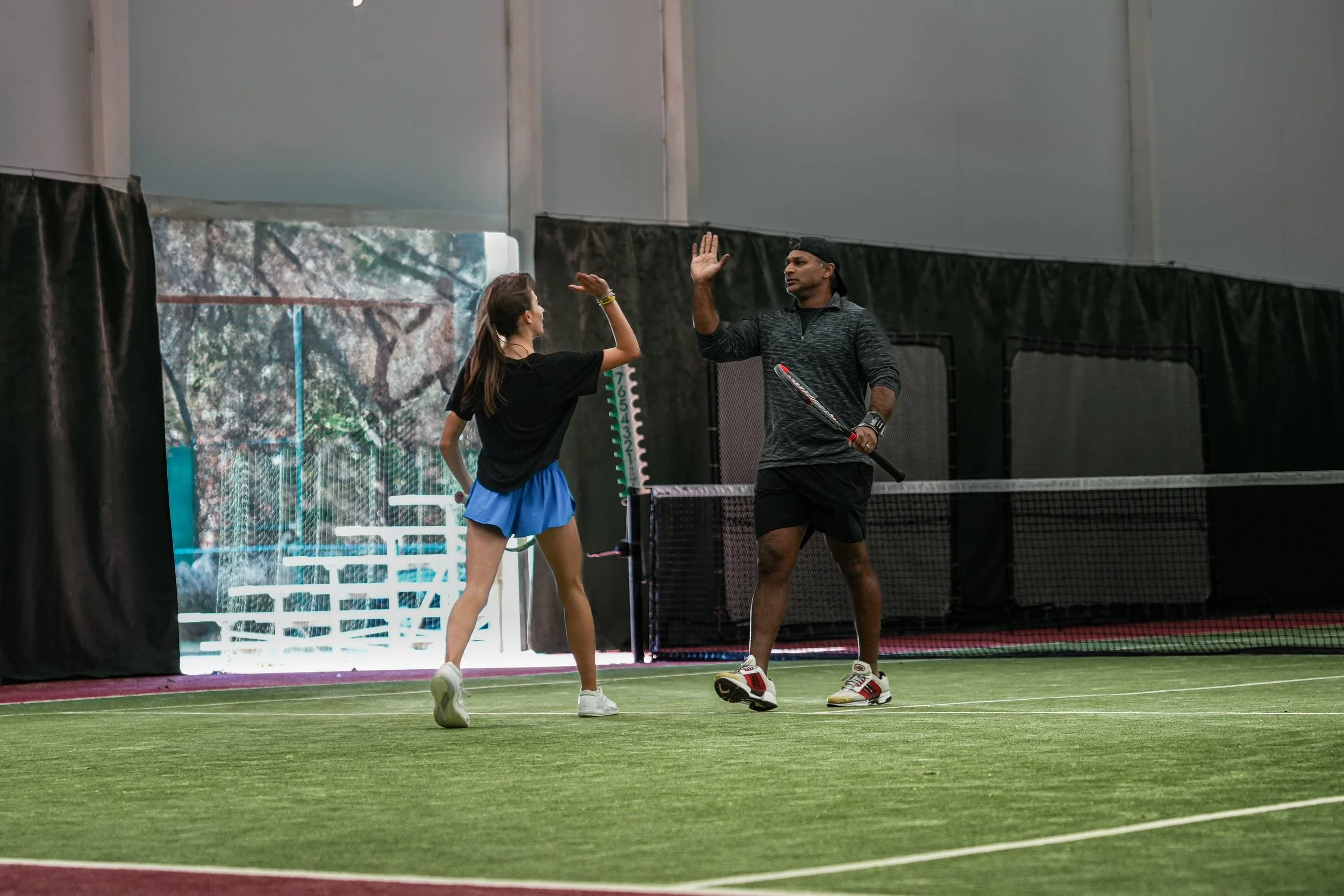 A young girl and an adult man playing tennis indoors, giving each other high-fives on a green tennis court.