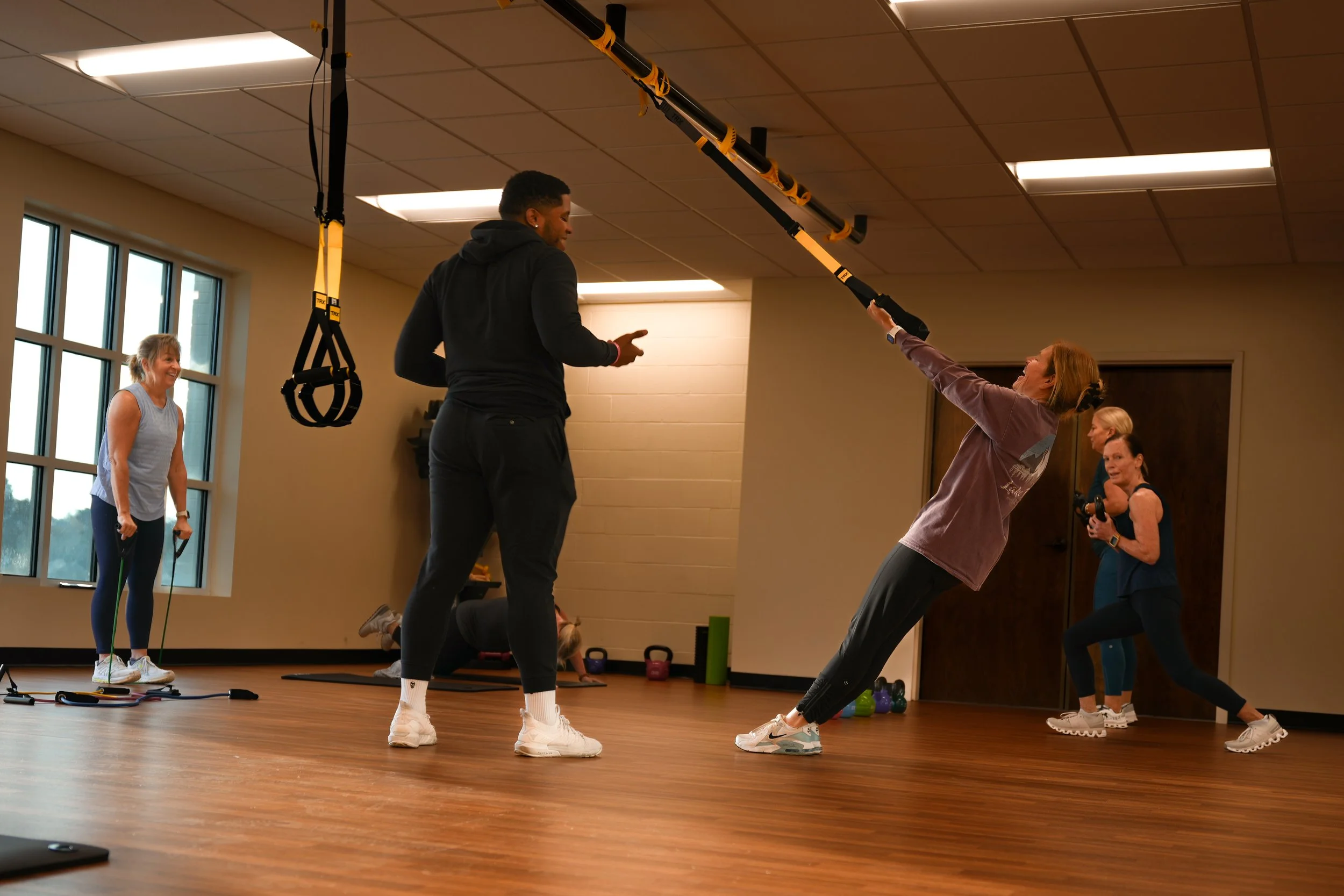 A fitness instructor guides a woman in a suspension training class, with other women exercising in the background in a well-lit gym with wood flooring.
