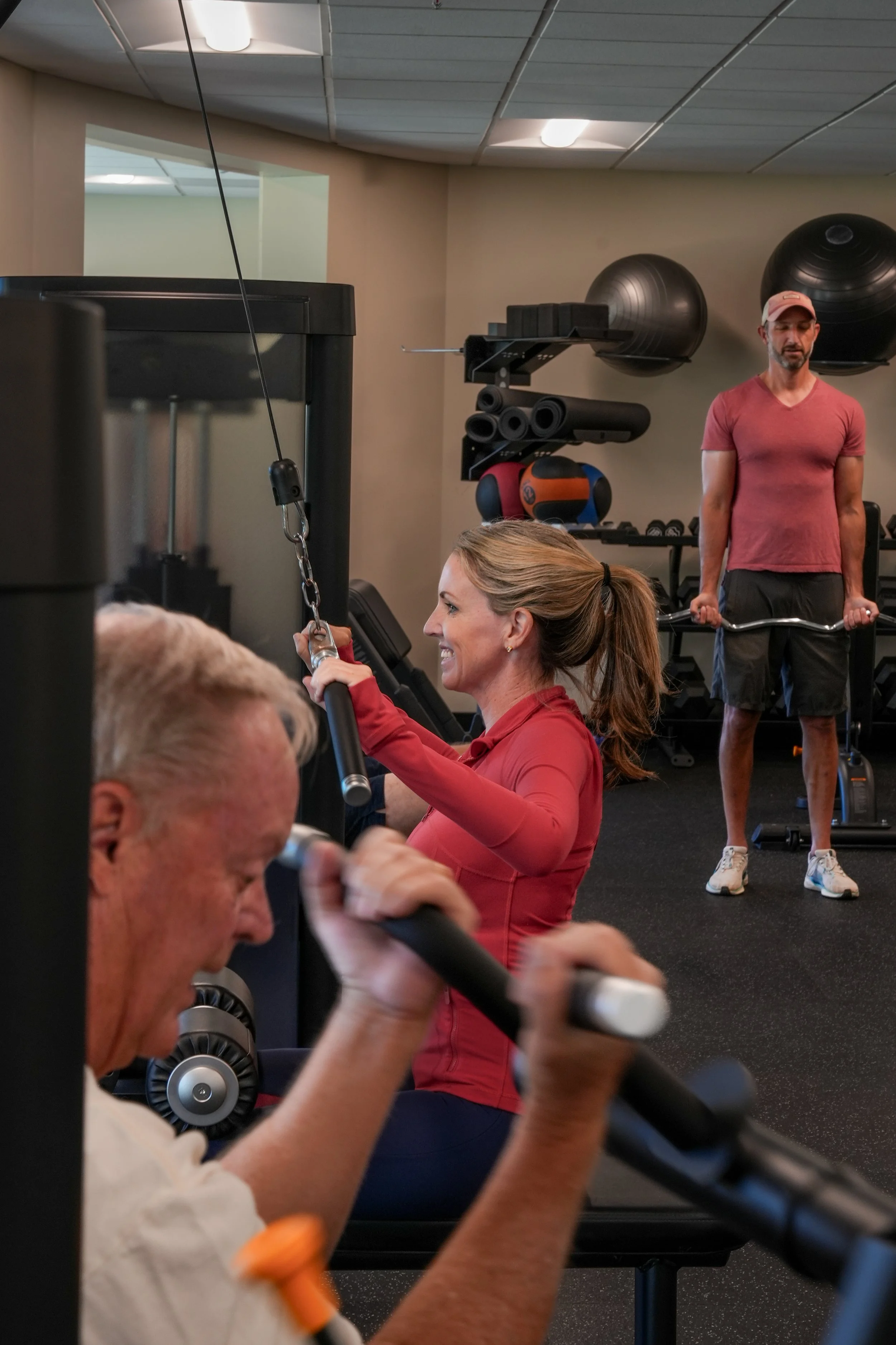 People exercising at a gym, including a woman using a cable machine, an elderly man lifting a barbell, and a man in the background doing bicep curls.