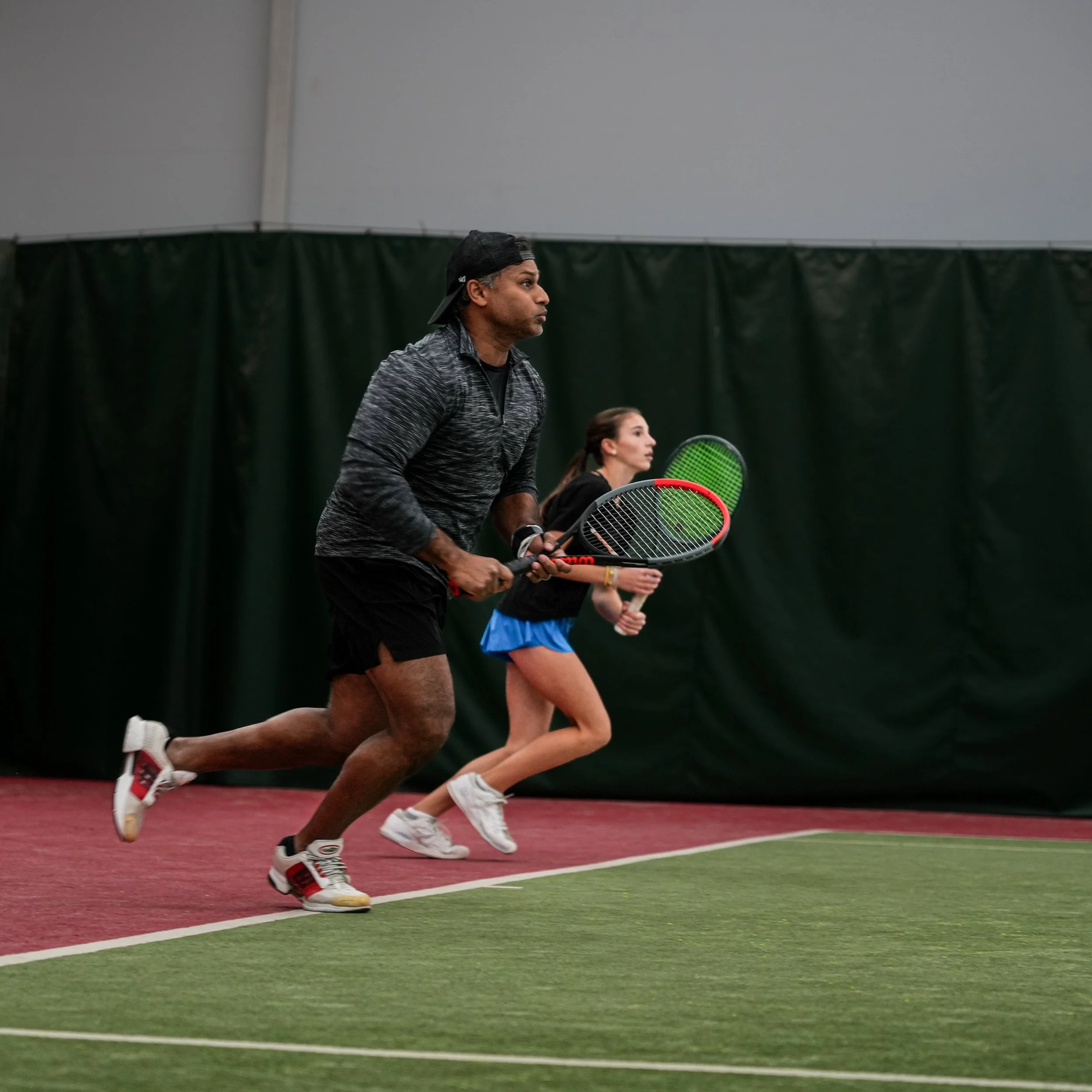 Two people playing tennis indoors, running on a court with green and red flooring, holding tennis rackets, with a dark curtain backdrop.