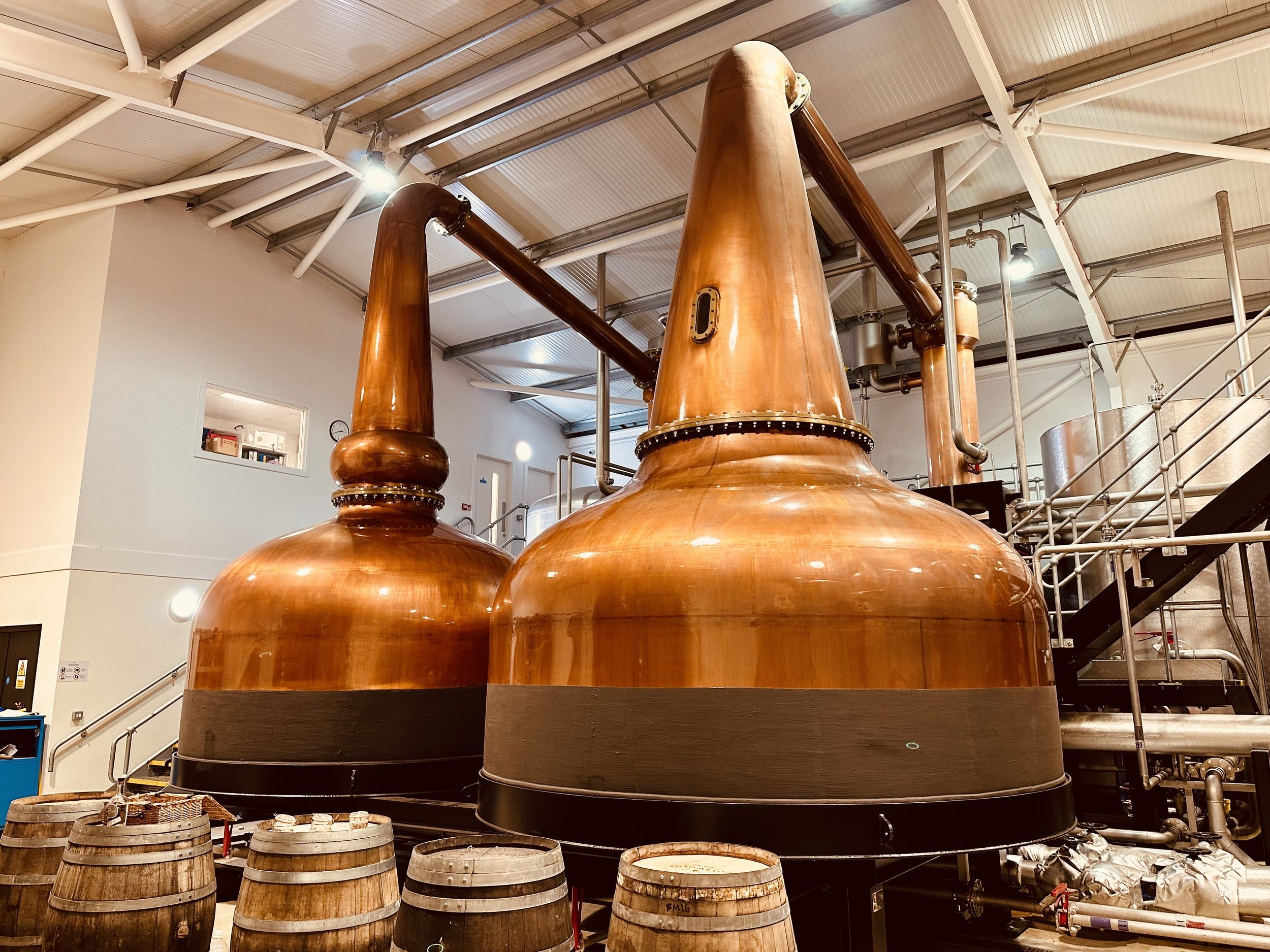A pair of large copper pot stills at The Cotswolds Distillery, with wooden casks in the foreground