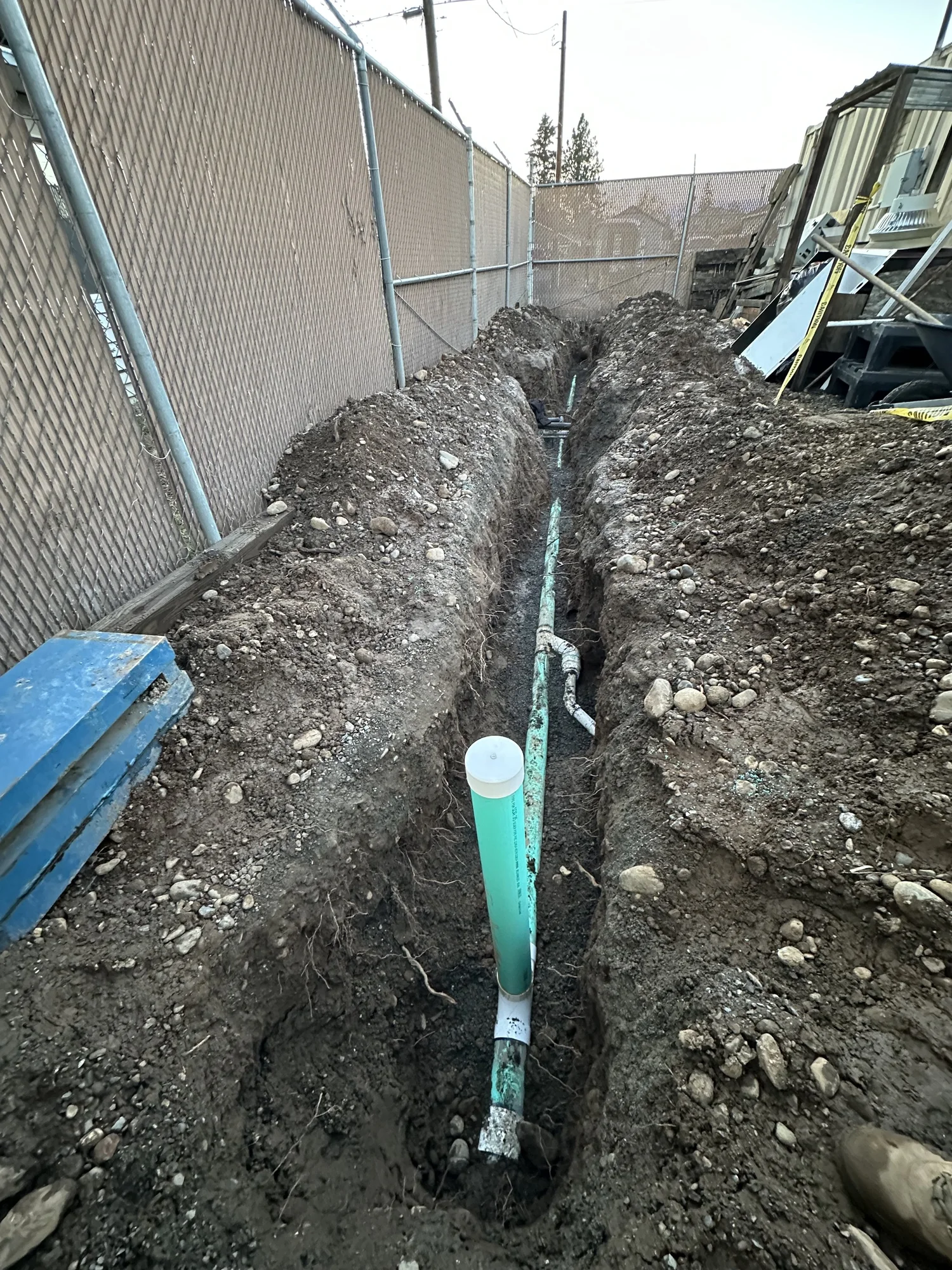 Sewer piping and a cleanout exposed in an excavated trench beside a building