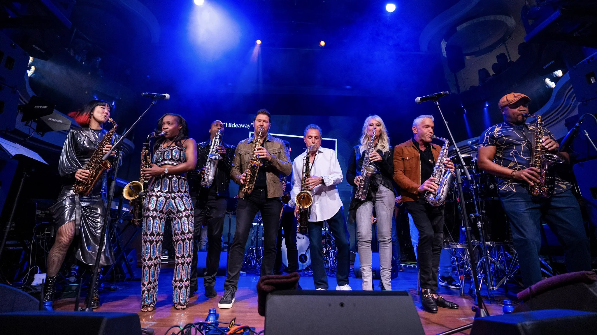 Musicians on stage with saxophones, standing in a row during a live performance in a concert hall with blue lighting.